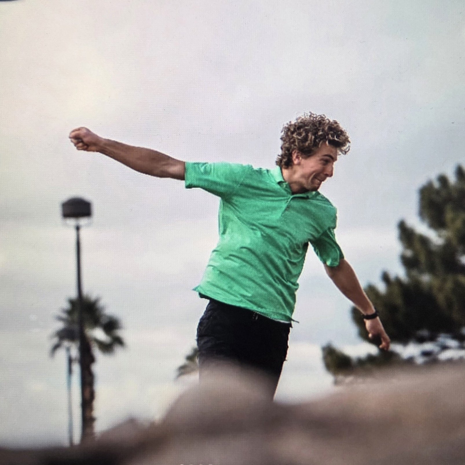 A man with curly hair in a green shirt and black shorts is jumping with his arms extended in an outdoor setting, with trees and a cloudy sky in the background.