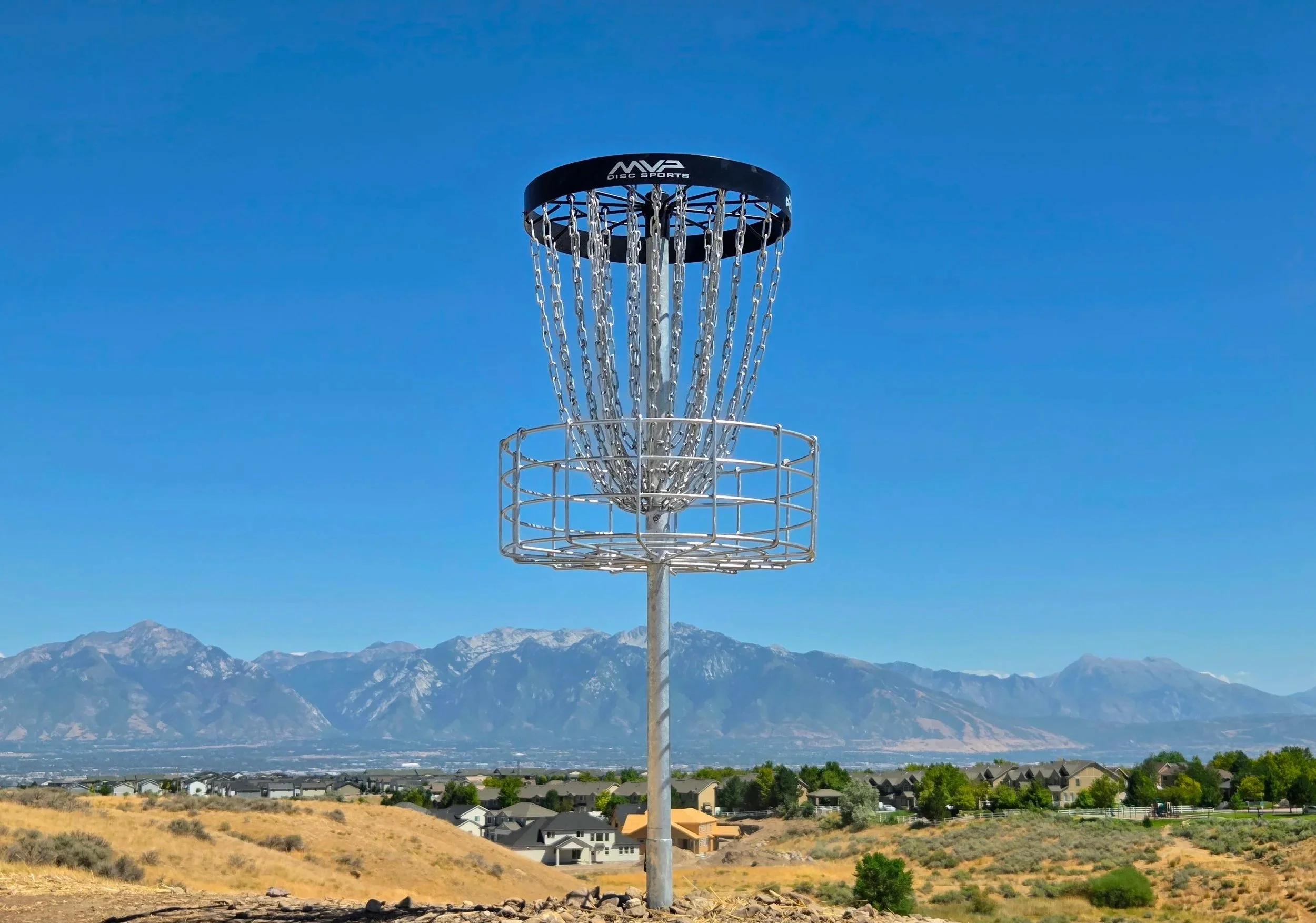 A disc golf basket stands outdoors against a mountain range and blue sky background.