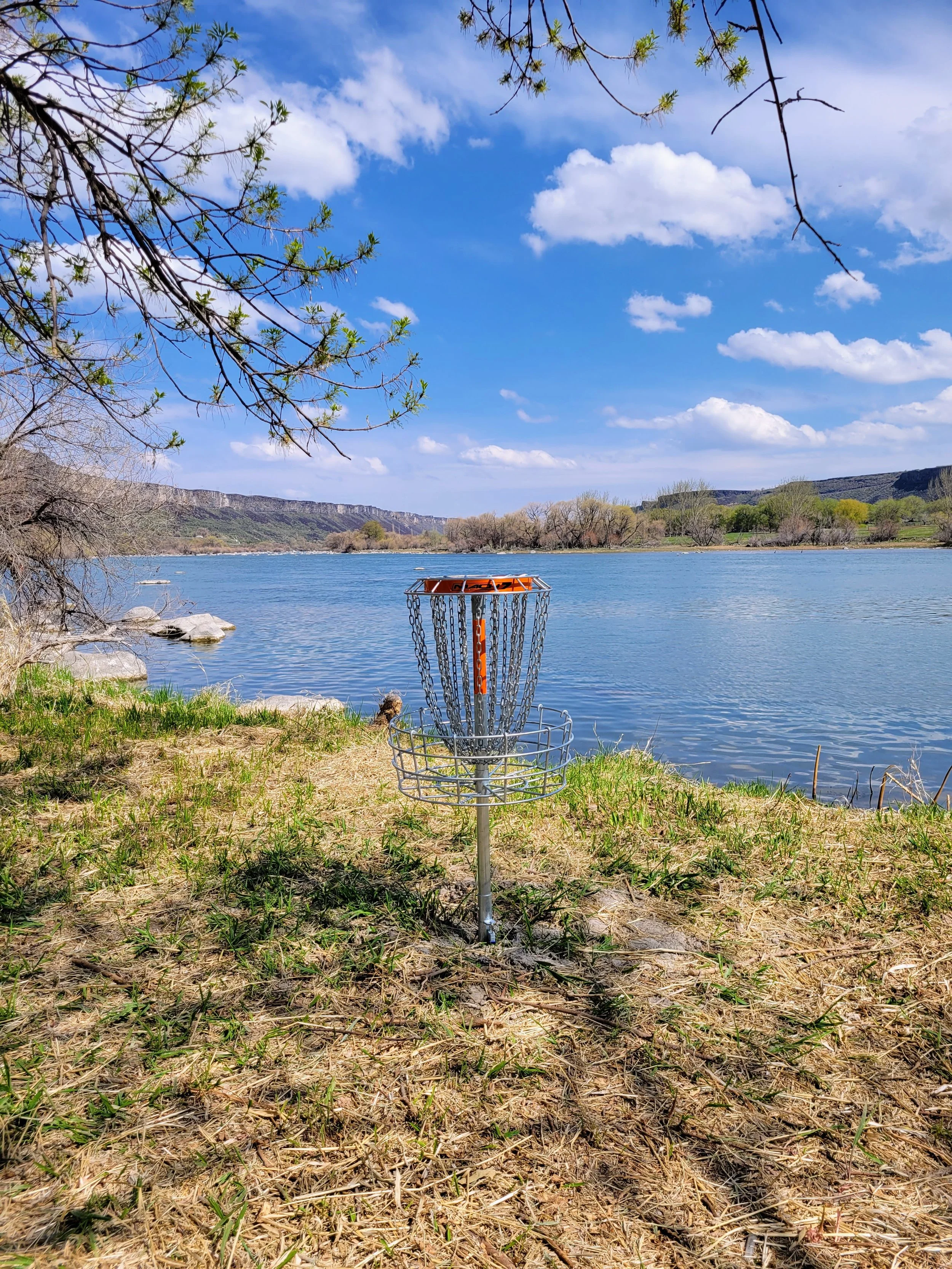 A disc golf basket on a grass shore by a lake with trees and hills in the background under a partly cloudy sky.