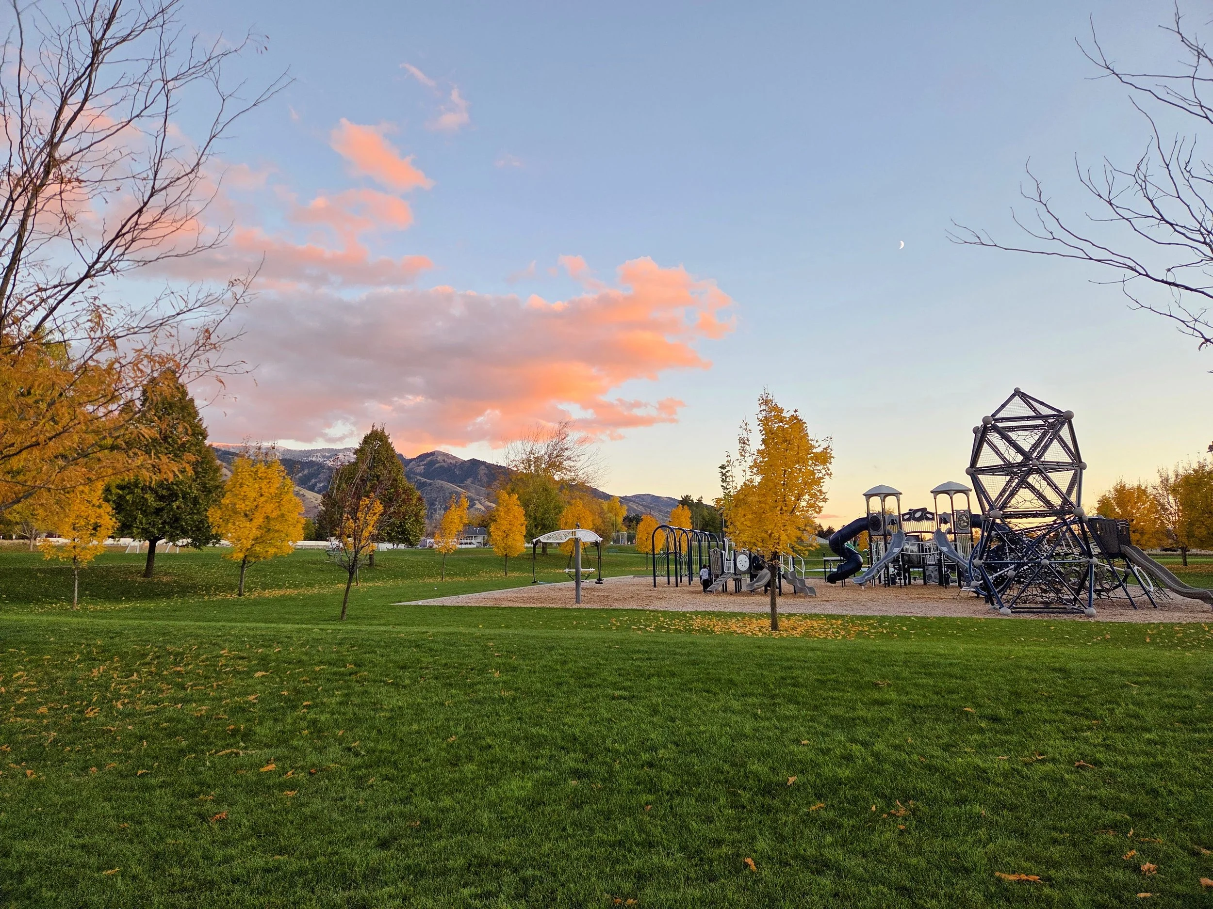 A park during sunset with colorful clouds, trees with orange and yellow leaves, and a mountain range in the background. There is a playground structure with slides and climbing equipment on the right side of the image.