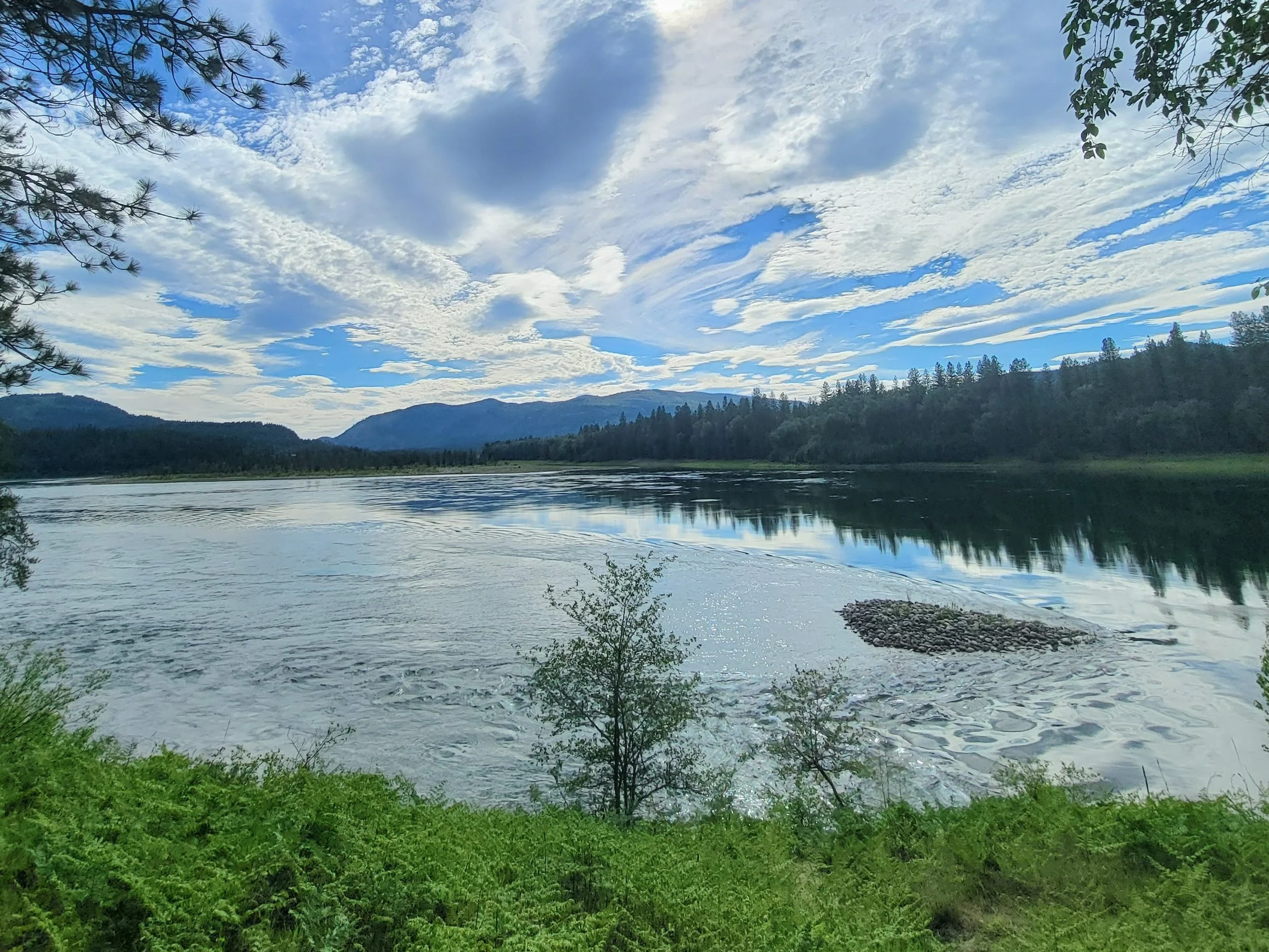 A scenic view of a large body of water surrounded by trees and hills, with a blue sky and scattered clouds above.