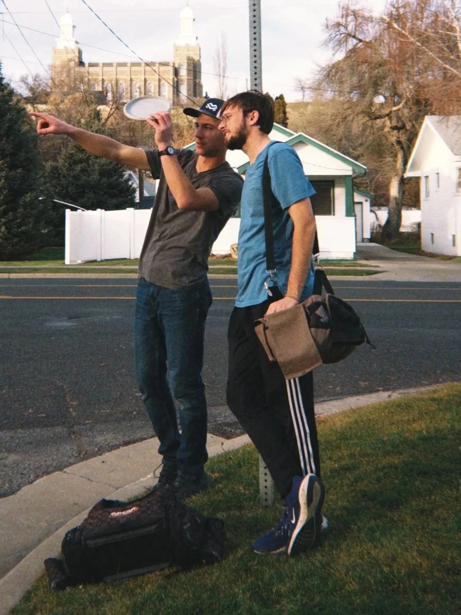 Two young men standing on the sidewalk near a signpost, one pointing while the other looks on. One man is wearing a black cap and gray t-shirt, the other is in a blue t-shirt with a backpack. There is a black bag on the ground beside them. The background features houses, trees, and a church or castle-like building.