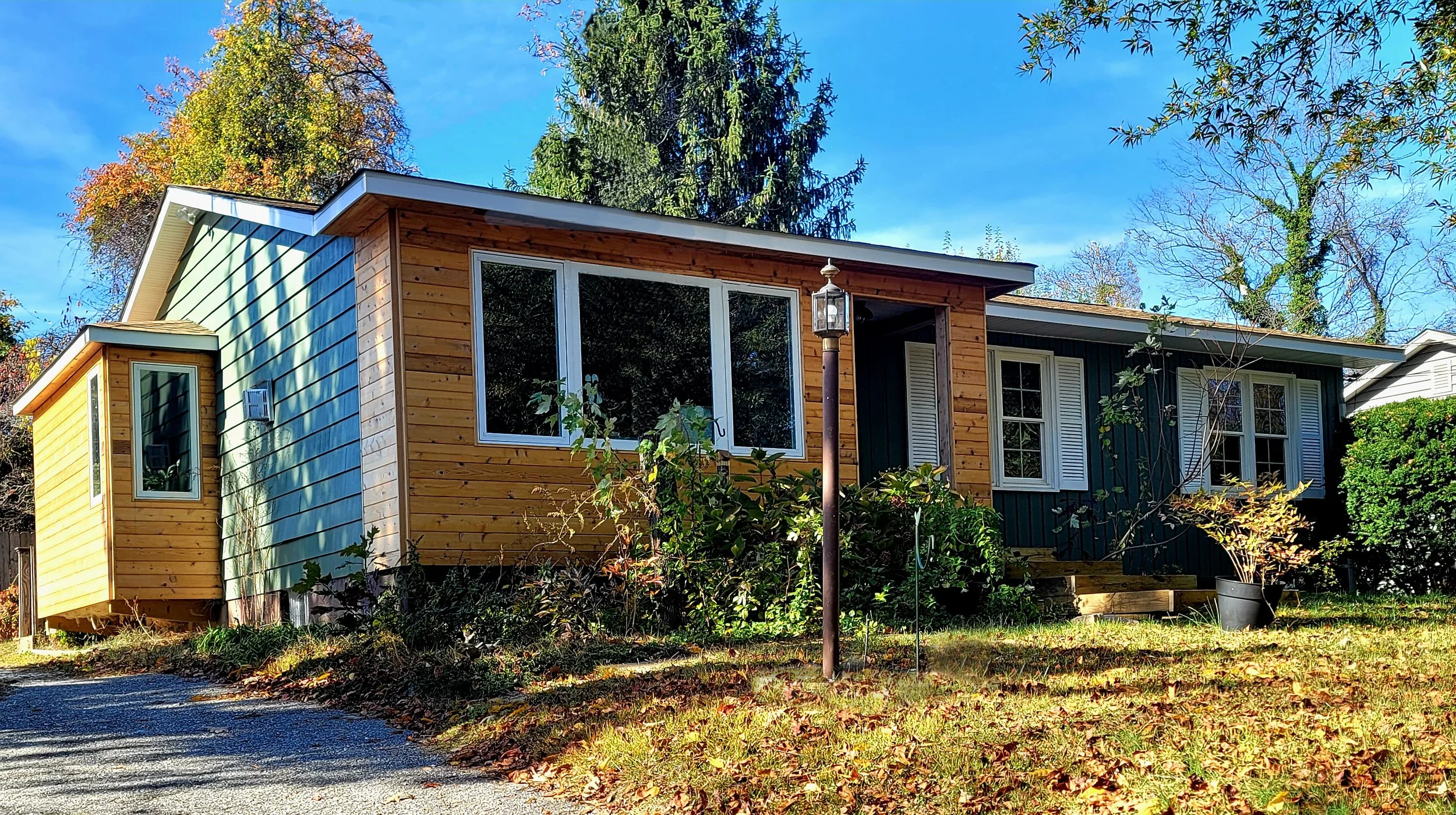 A house with mixed siding, one side with green shingles and the other with natural wood, surrounded by trees and a lawn with fallen leaves, under a clear blue sky.