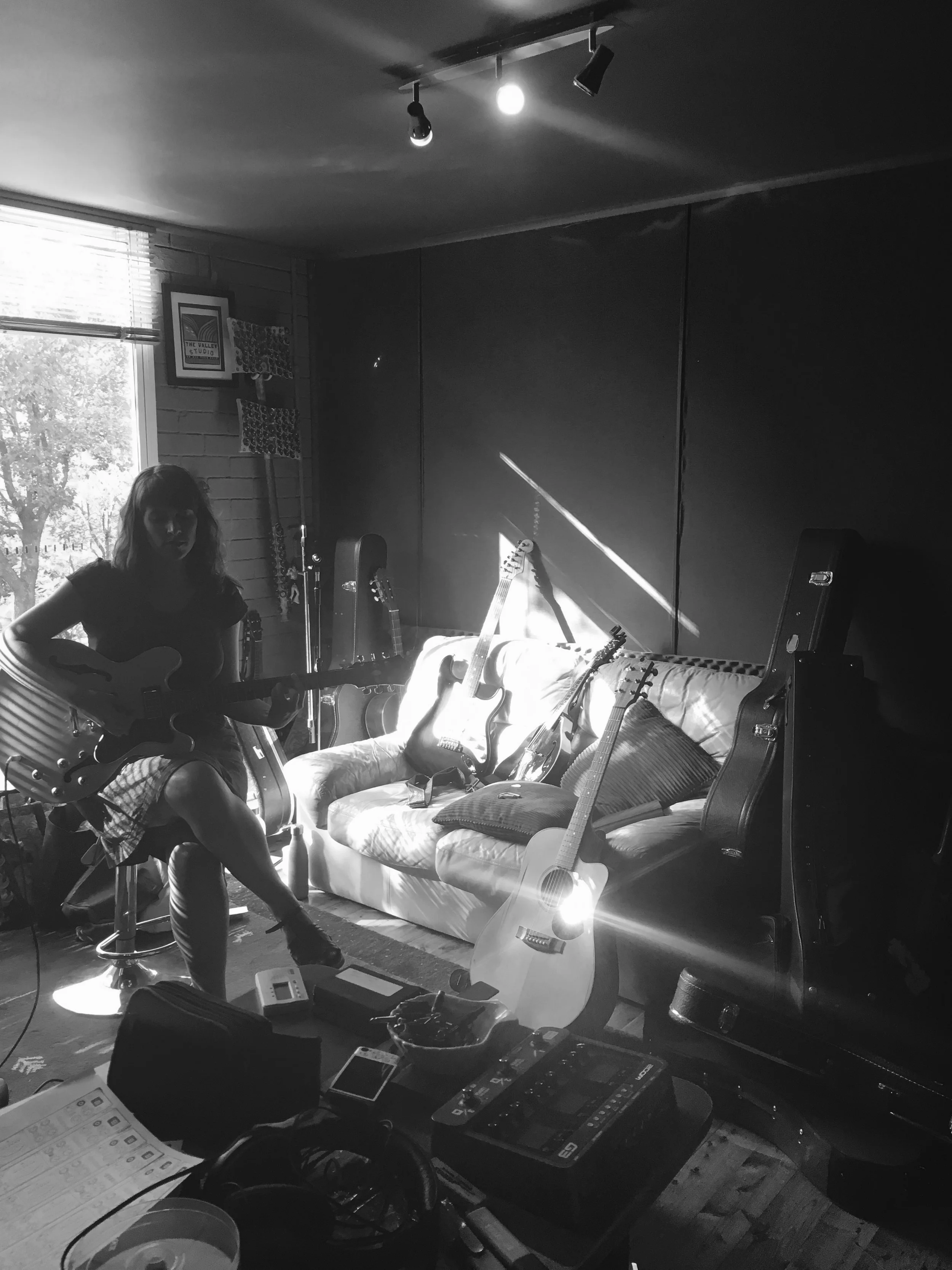 A woman sitting on a stool playing an electric guitar in a music studio, with other guitars leaning against the wall, a sofa, and various music equipment.