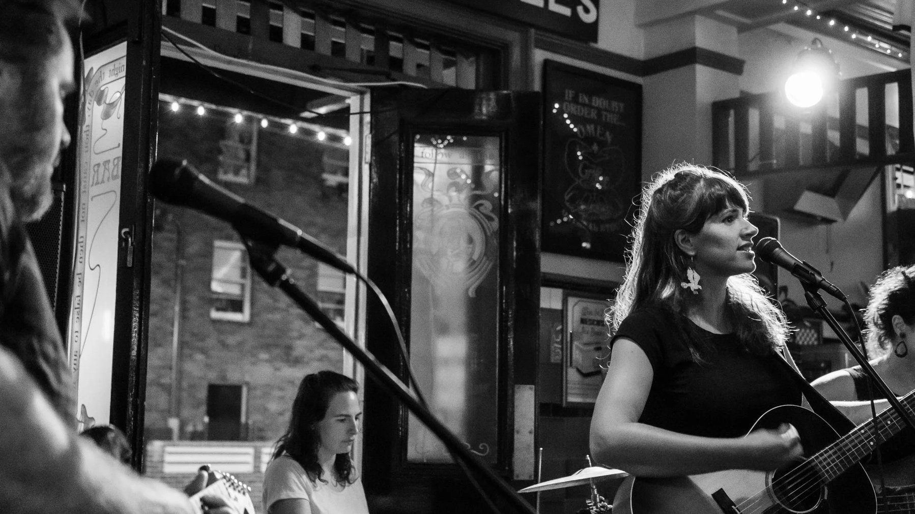A black and white photo of a woman singing and playing a guitar at a bar or cafe, with other musicians in the background and posters on the walls.