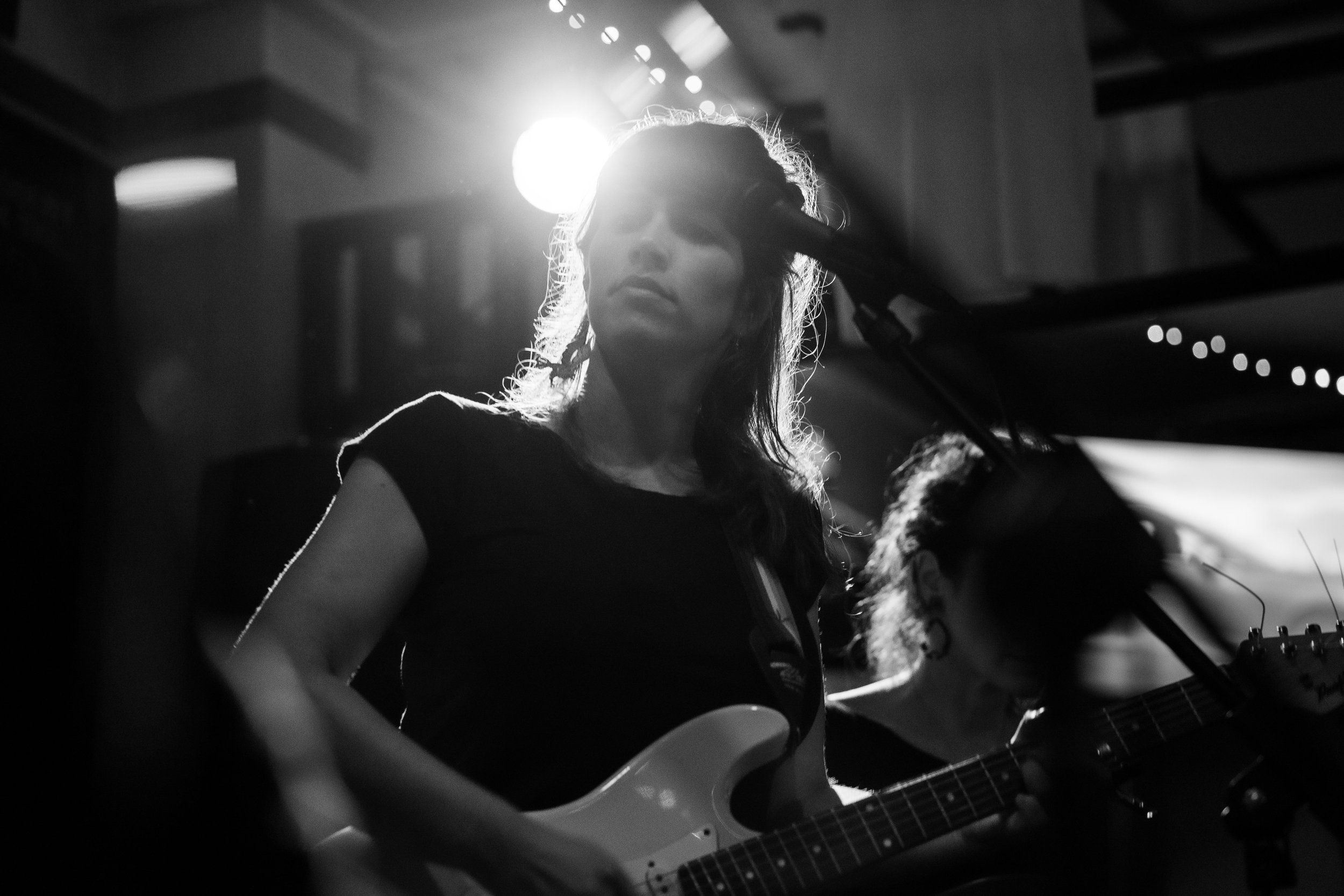 A woman playing an electric guitar during a live performance, illuminated by stage lighting with a bright light source behind her.