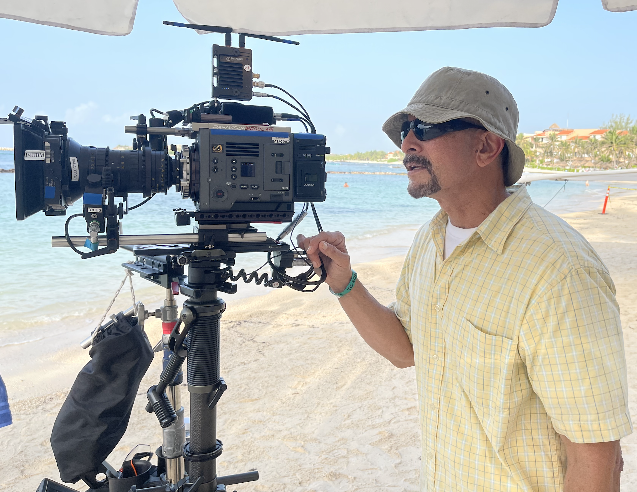 Man operating a professional video camera on a beach, wearing a beige bucket hat, sunglasses, and a yellow checkered shirt.