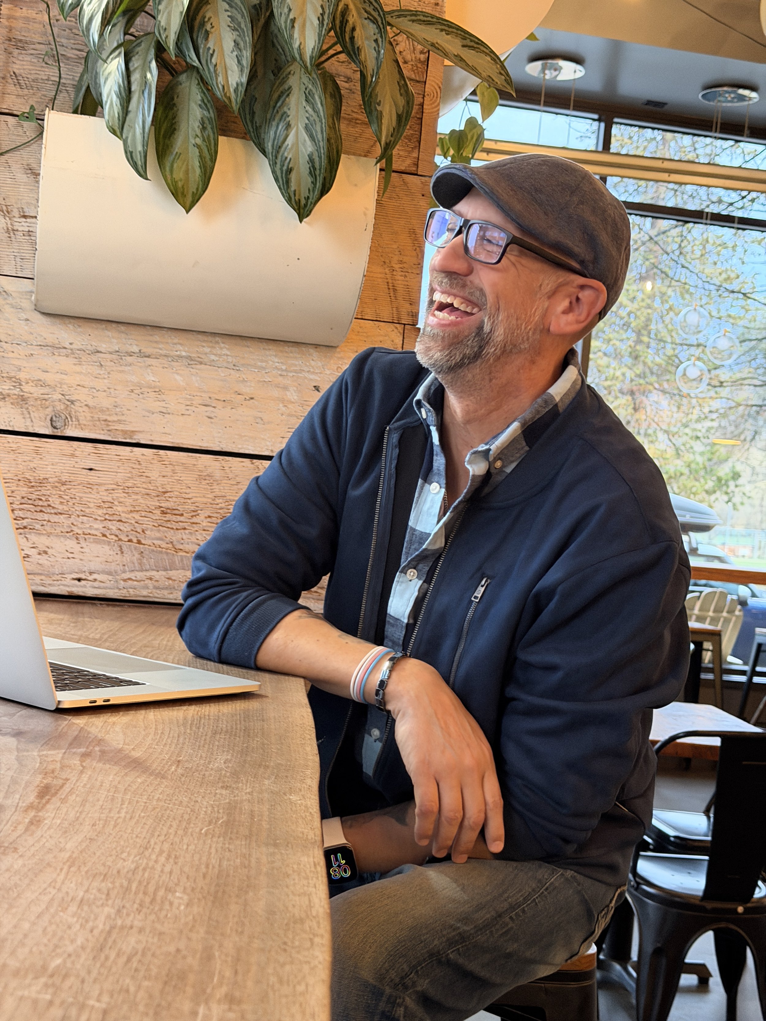 A man with glasses and a flat cap sitting at a wooden table, smiling and looking to his right, in a cafe or restaurant with a window showing trees outside.