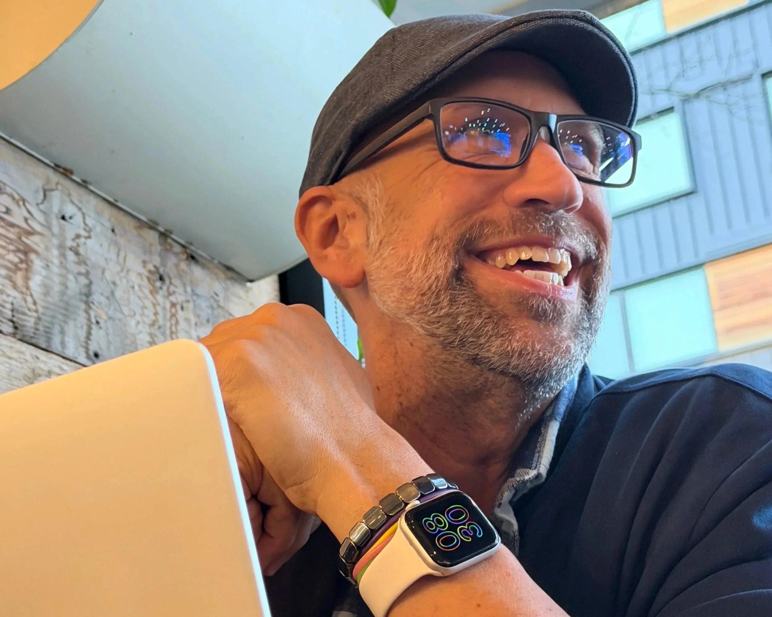 A smiling middle-aged man in glasses and a gray cap, wearing a smartwatch and a bracelet, sitting in a cafe with a wooden wall and large windows in the background.
