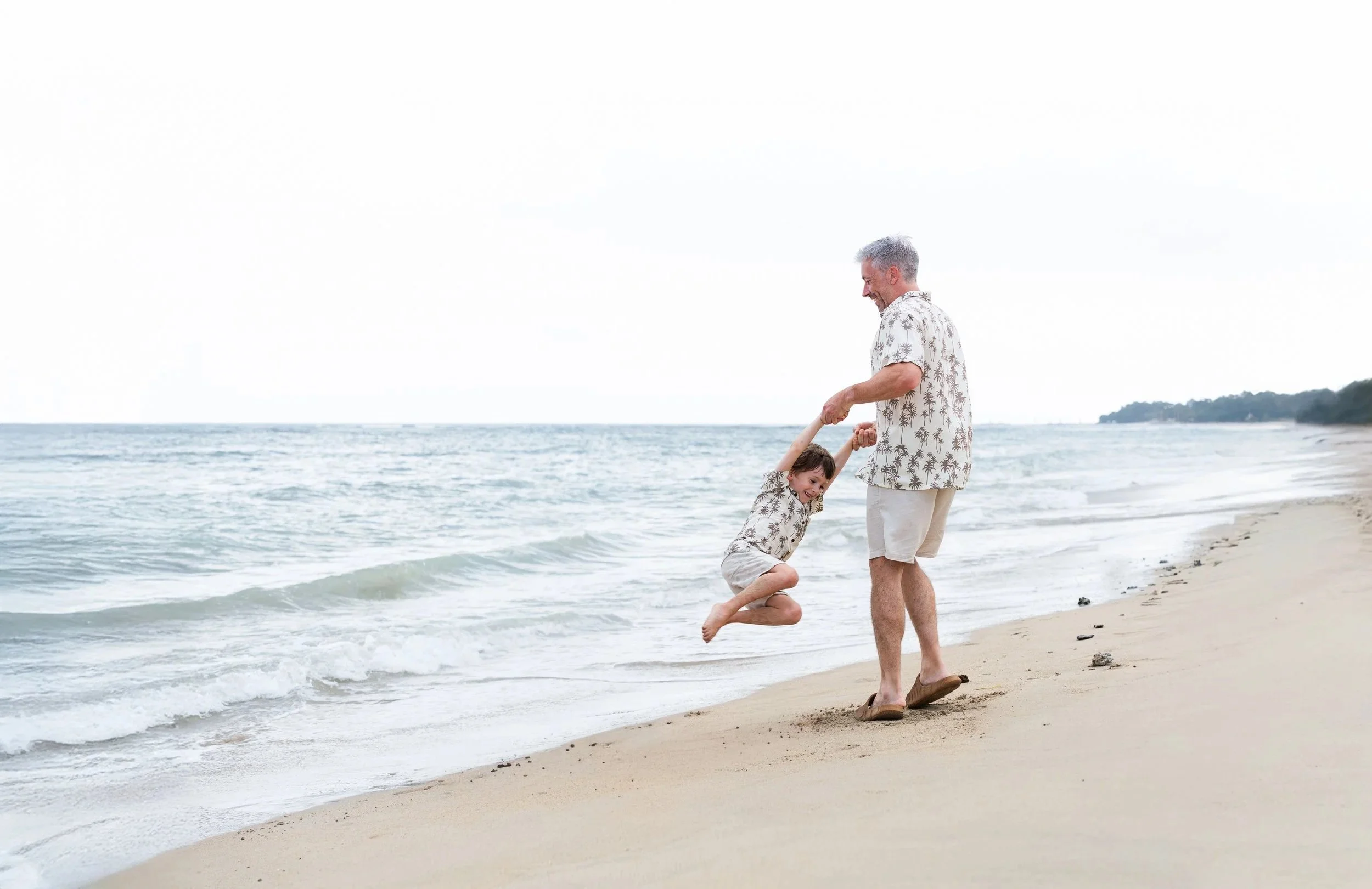 Een man en een kind spelen samen op het strand, terwijl de man het kind optilt boven de grond. Ze dragen matching Hawaiiaanse shirts en lijken plezier te hebben.