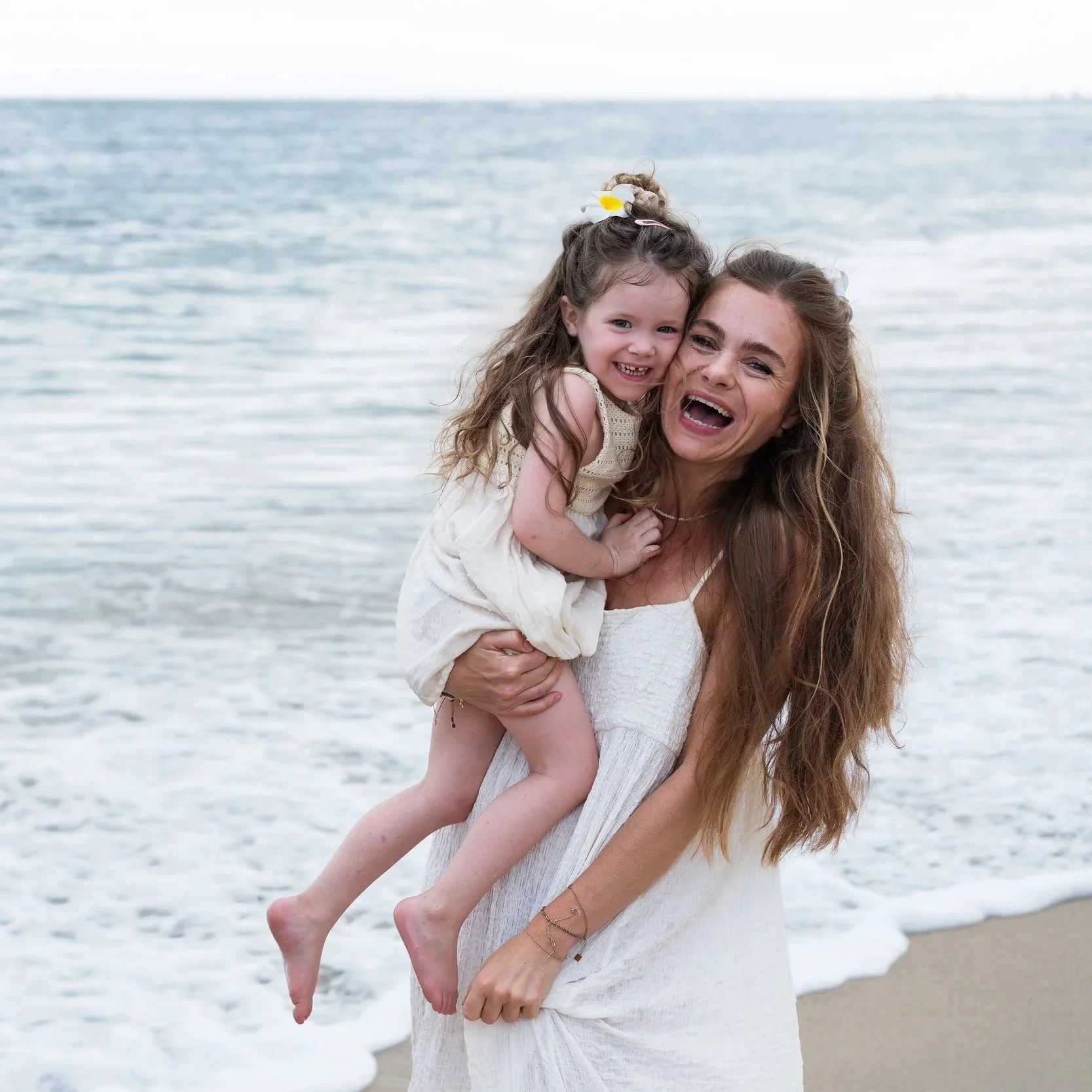 Vrouw houdt jonge meisje vast op strand, beide lachen, oceaan op achtergrond
