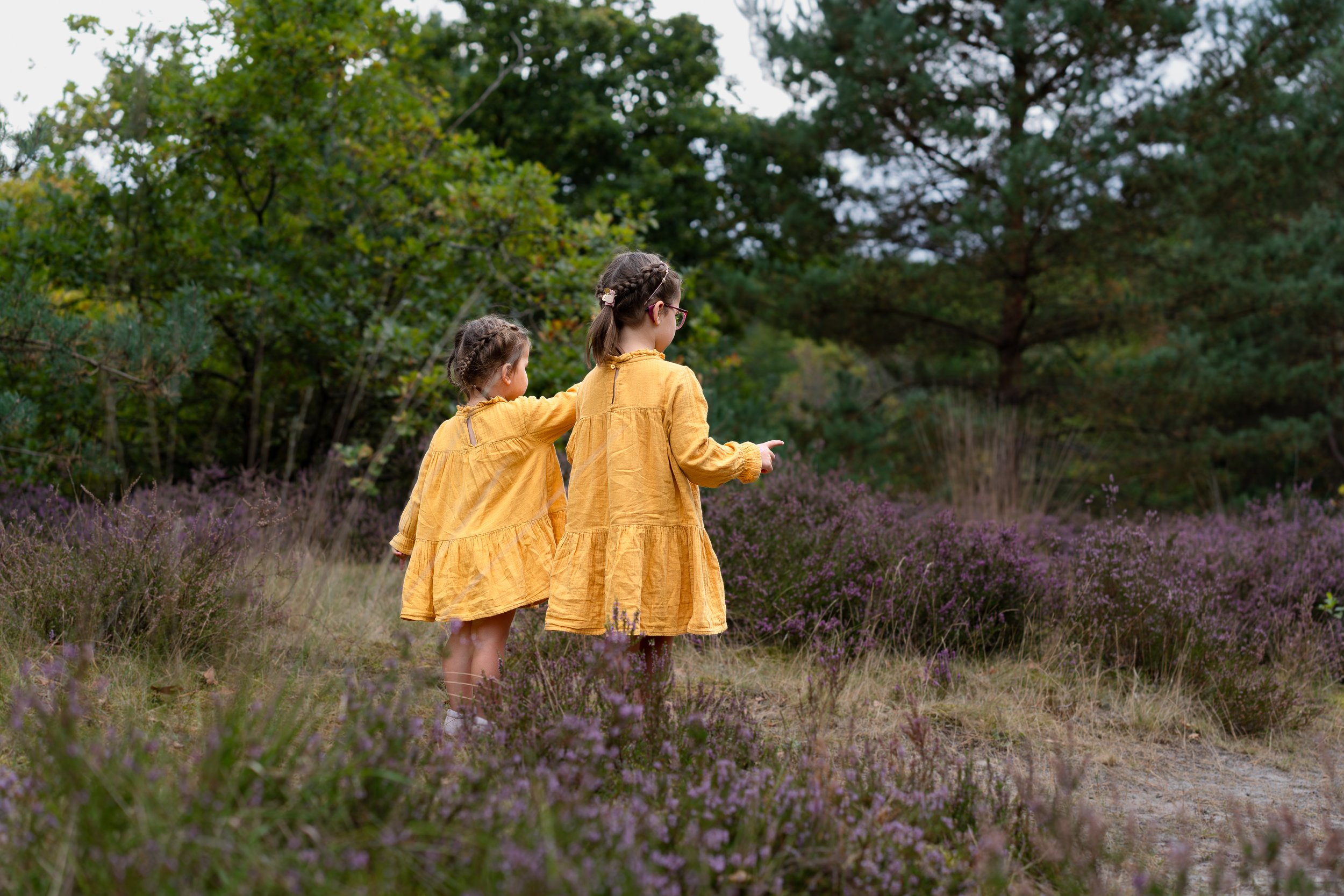 Twee jonge meisjes in gele jurken wandelen door een veld met paarse bloemen, omringd door groene bomen.