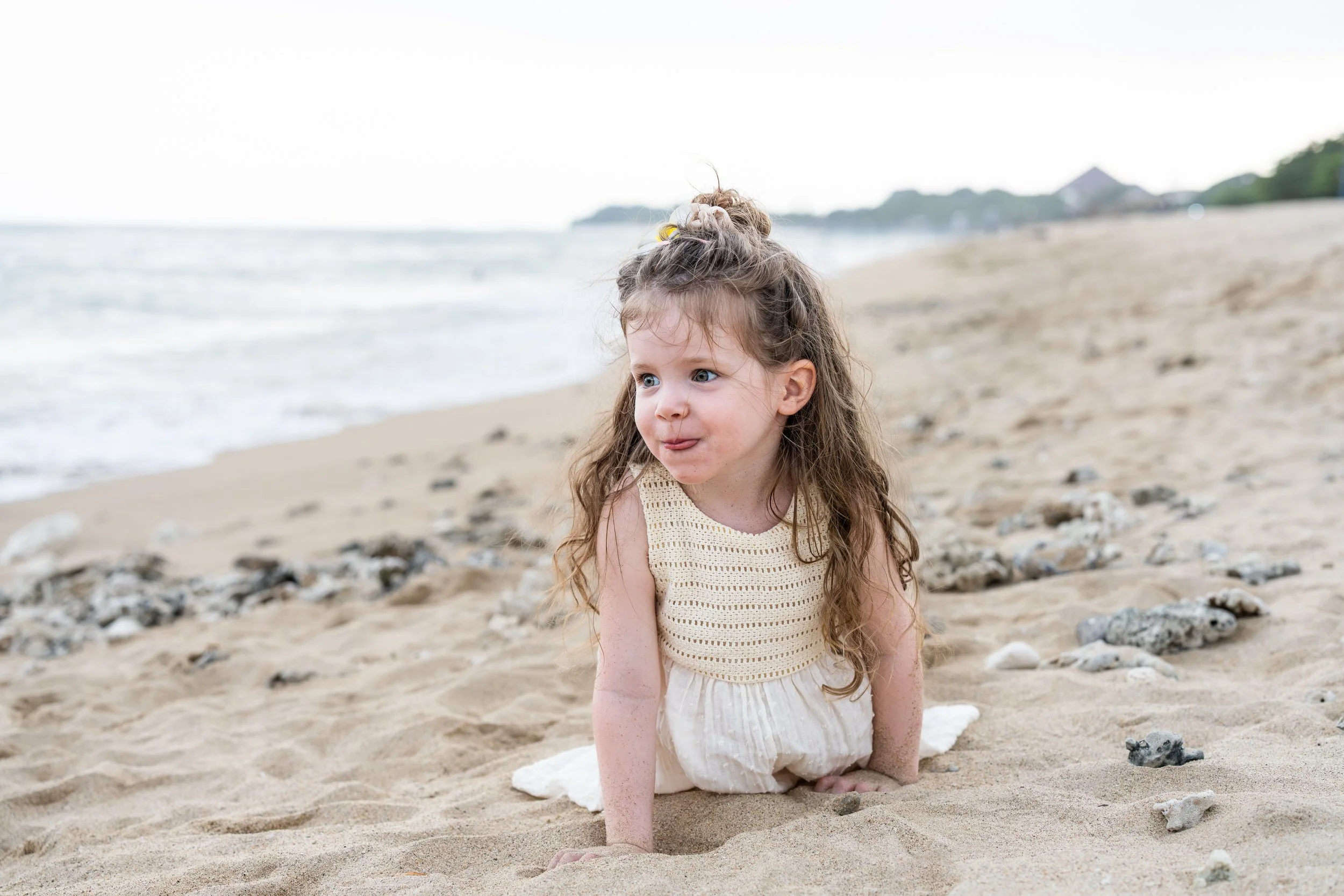 Een jonge meisje met lang, krullend haar zit op het zand bij het strand, gekleed in een lichte, gebreide top en een lichtkleurige broek, met de zee en de hemel op de achtergrond.