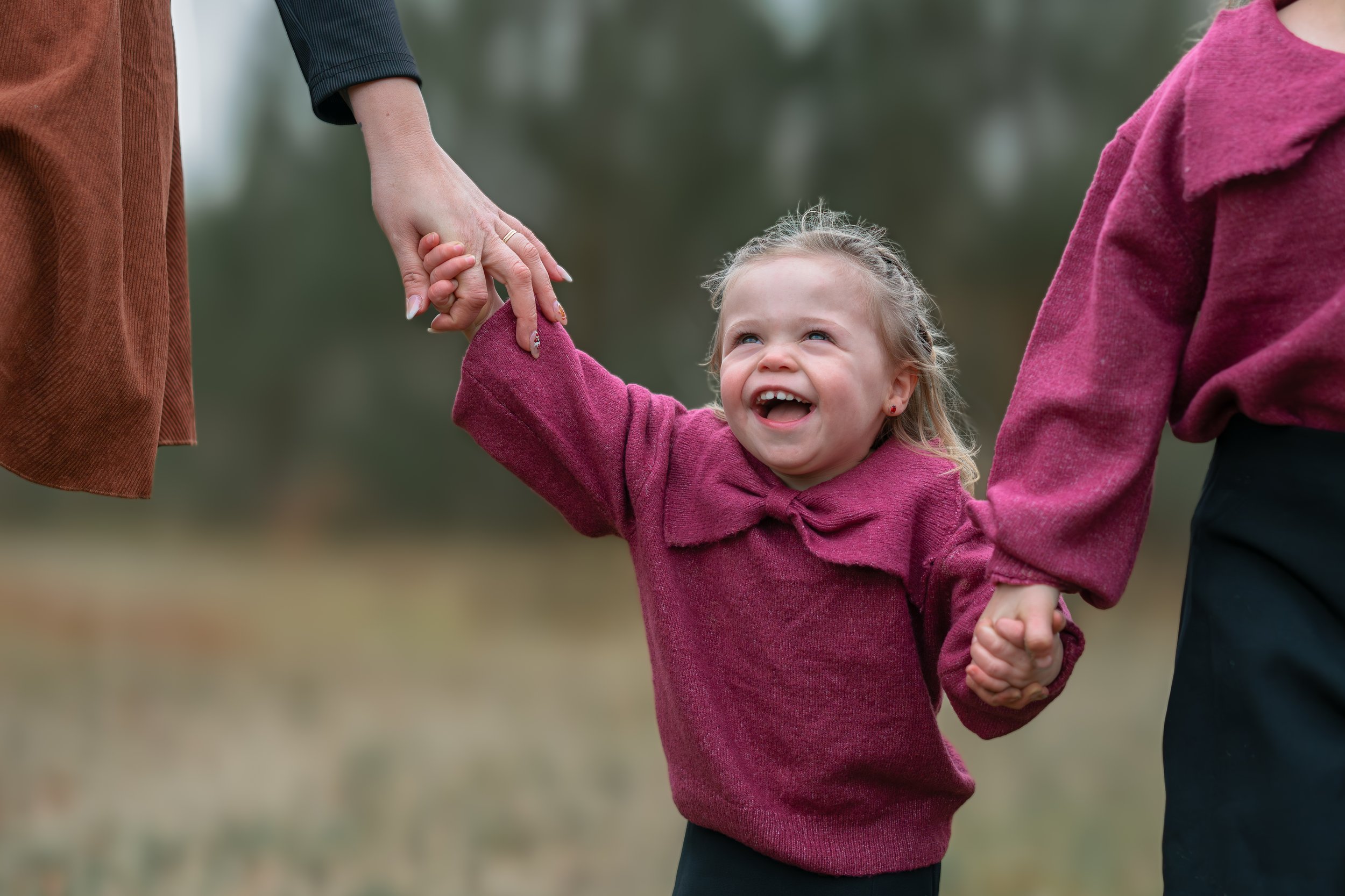 Een jonge meisje die lachend wordt vastgehouden door twee volwassenen in een veld, met een glimlach en stralende ogen.