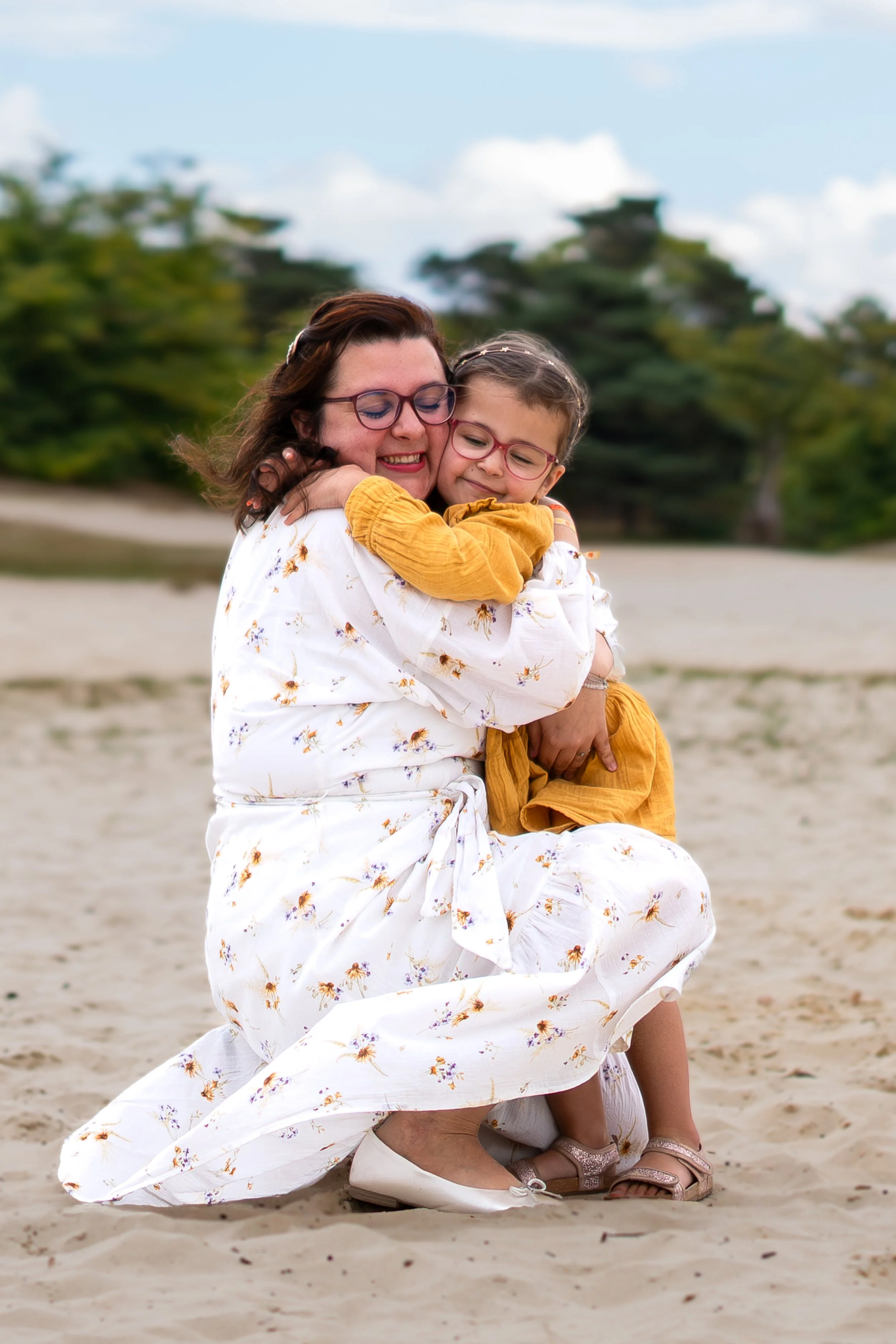Twee vrouwen omarmen elkaar op het strand, lachend en knuffelend, met groene bomen en een blauwe lucht op de achtergrond.