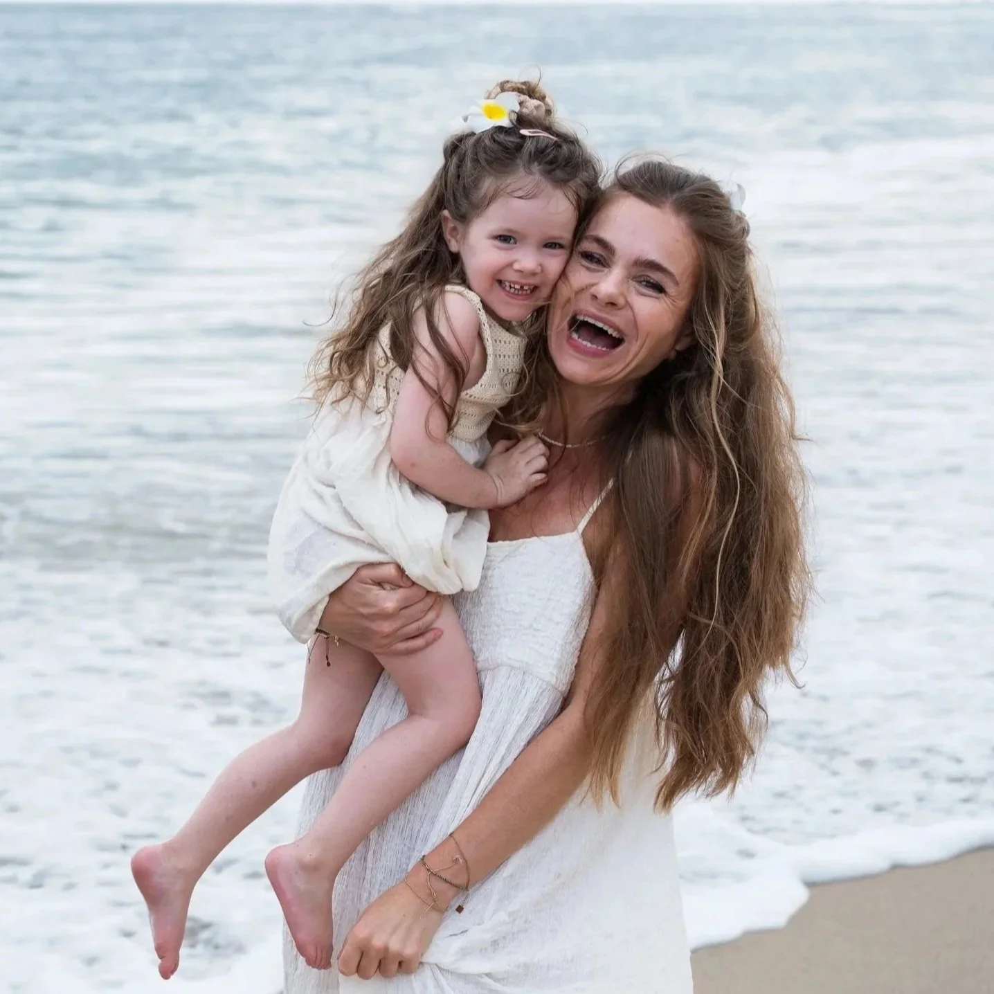 Vrouw die een lachende jonge meisje op haar schouder houdt aan het strand, met water op de achtergrond.