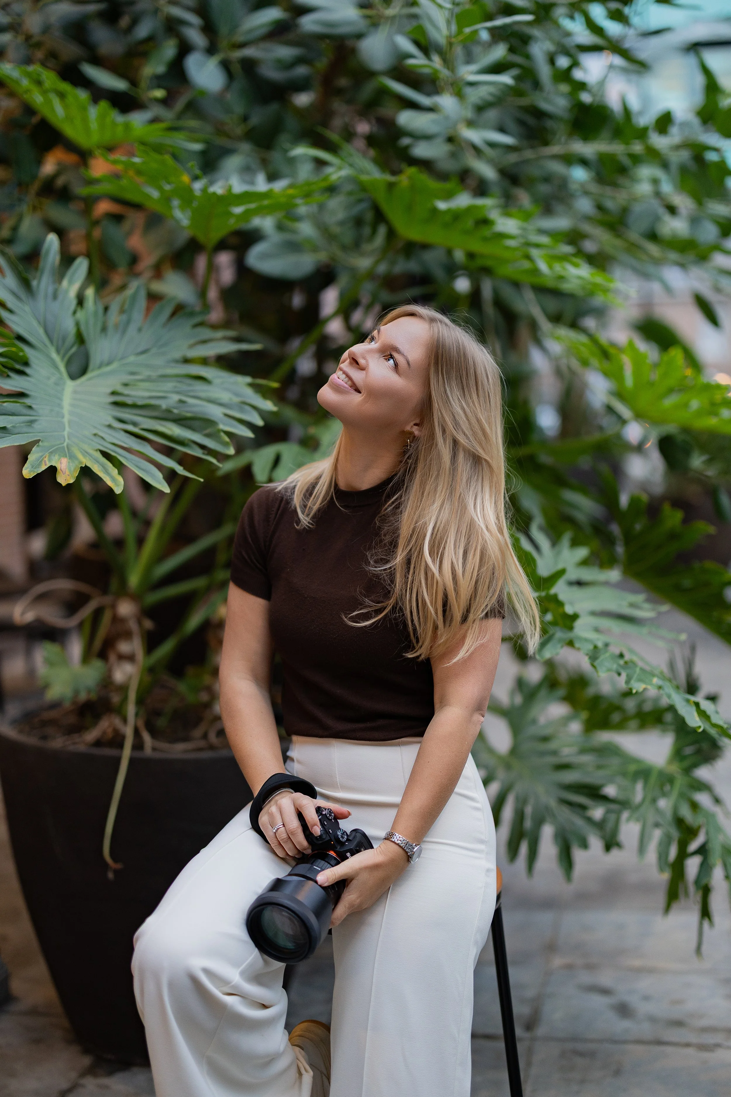 Een vrouw met lang blond haar, gekleed in een zwart t-shirt en witte broek, zit op een krukje in een ruimte met grote groene planten. Ze houdt een professionele camera met telelens in haar handen en kijkt omhoog met een lichte glimlach.