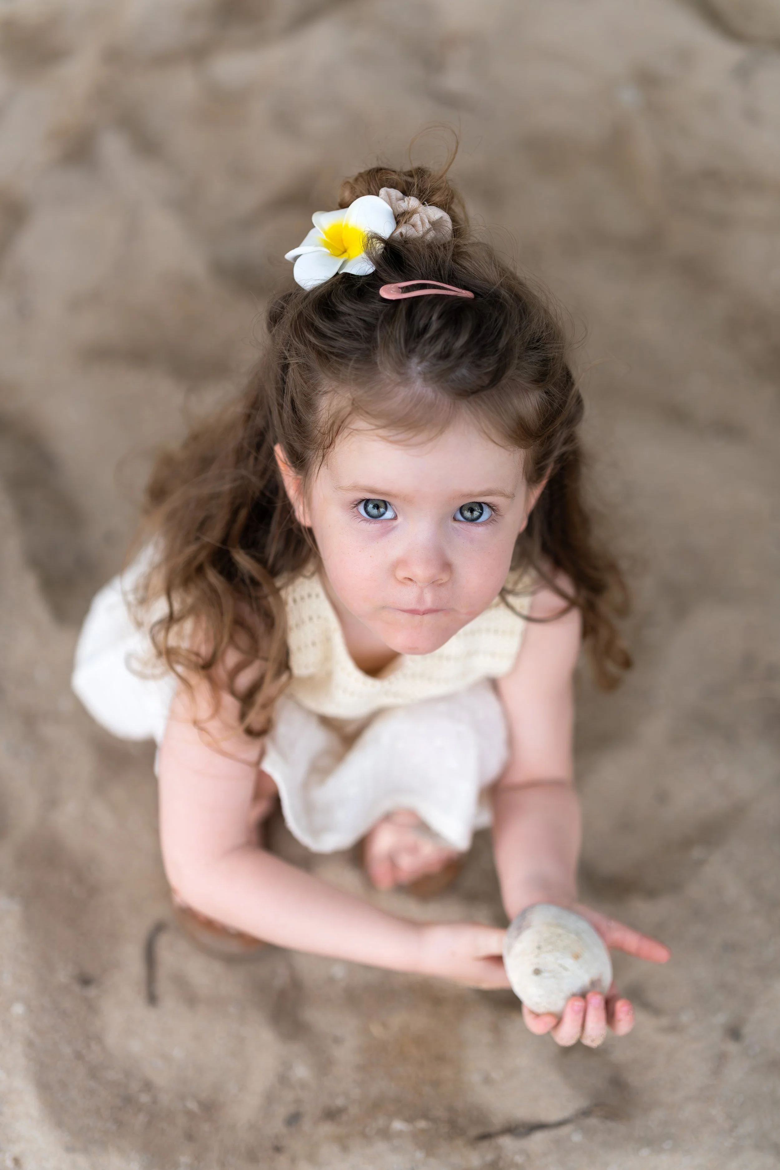 Kleine meisje met blauwe ogen, lange bruin haar met een bloem en haarspeld, zittend op zand terwijl ze een steen vasthoudt, aan een strand of zandbak.