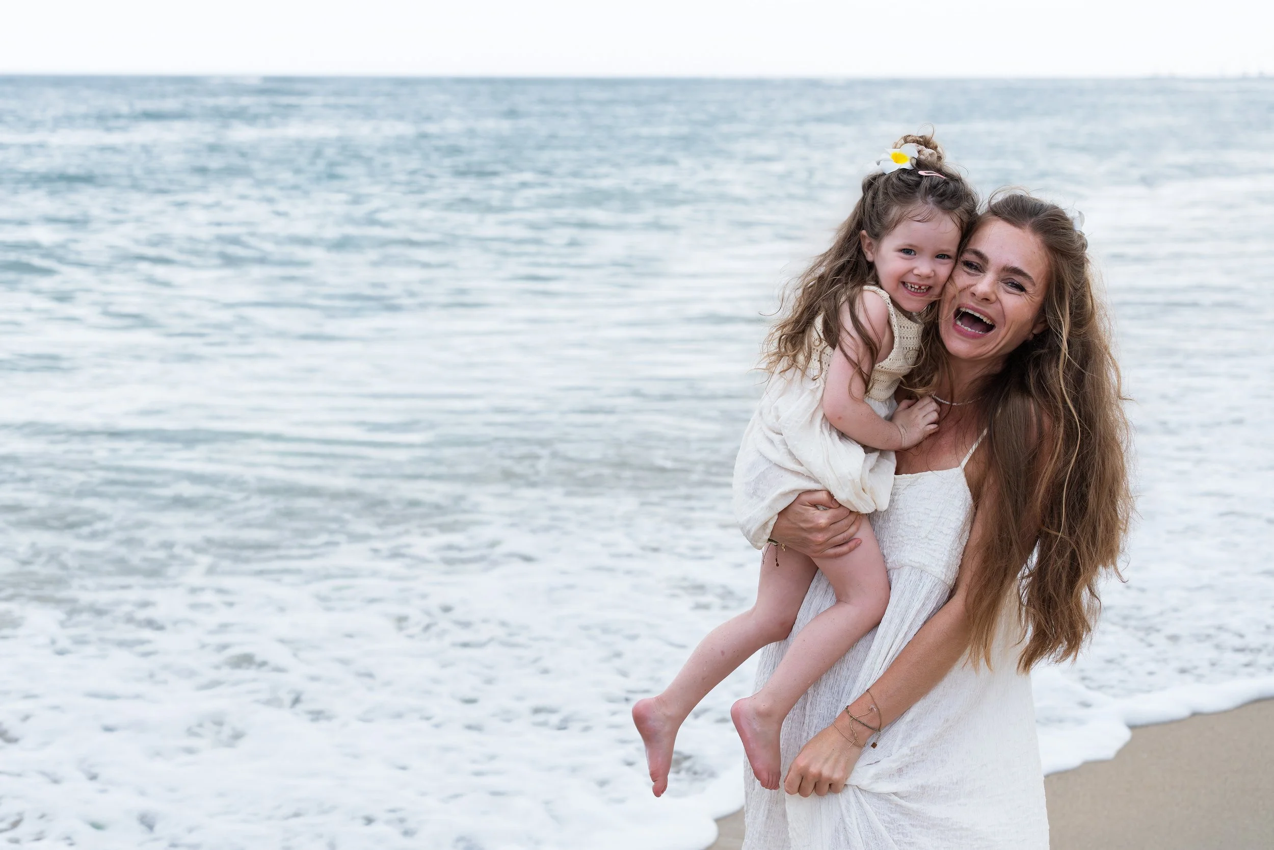 Vrouw houdt jonge meisje vast op strand, beide lachen, oceaan op achtergrond