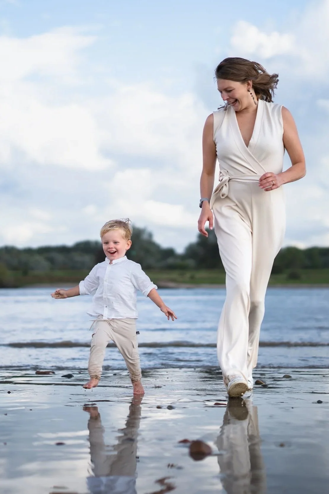 Vrouw en jongen lopen over het strand, plezier en lachen, met water en wolken op de achtergrond.