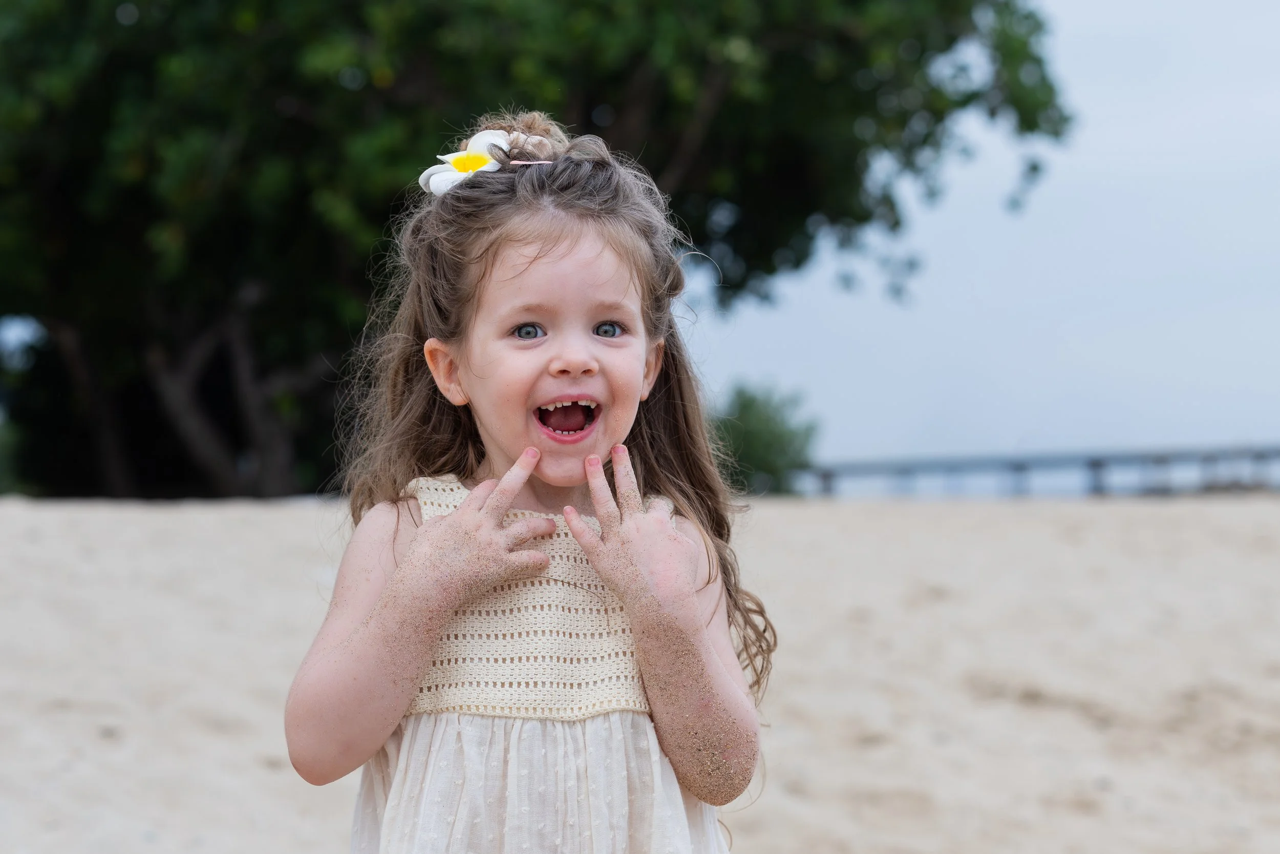 Lachende jonge meisje met zand op haar armen en gezicht, op het strand met bomen en een brug op de achtergrond.