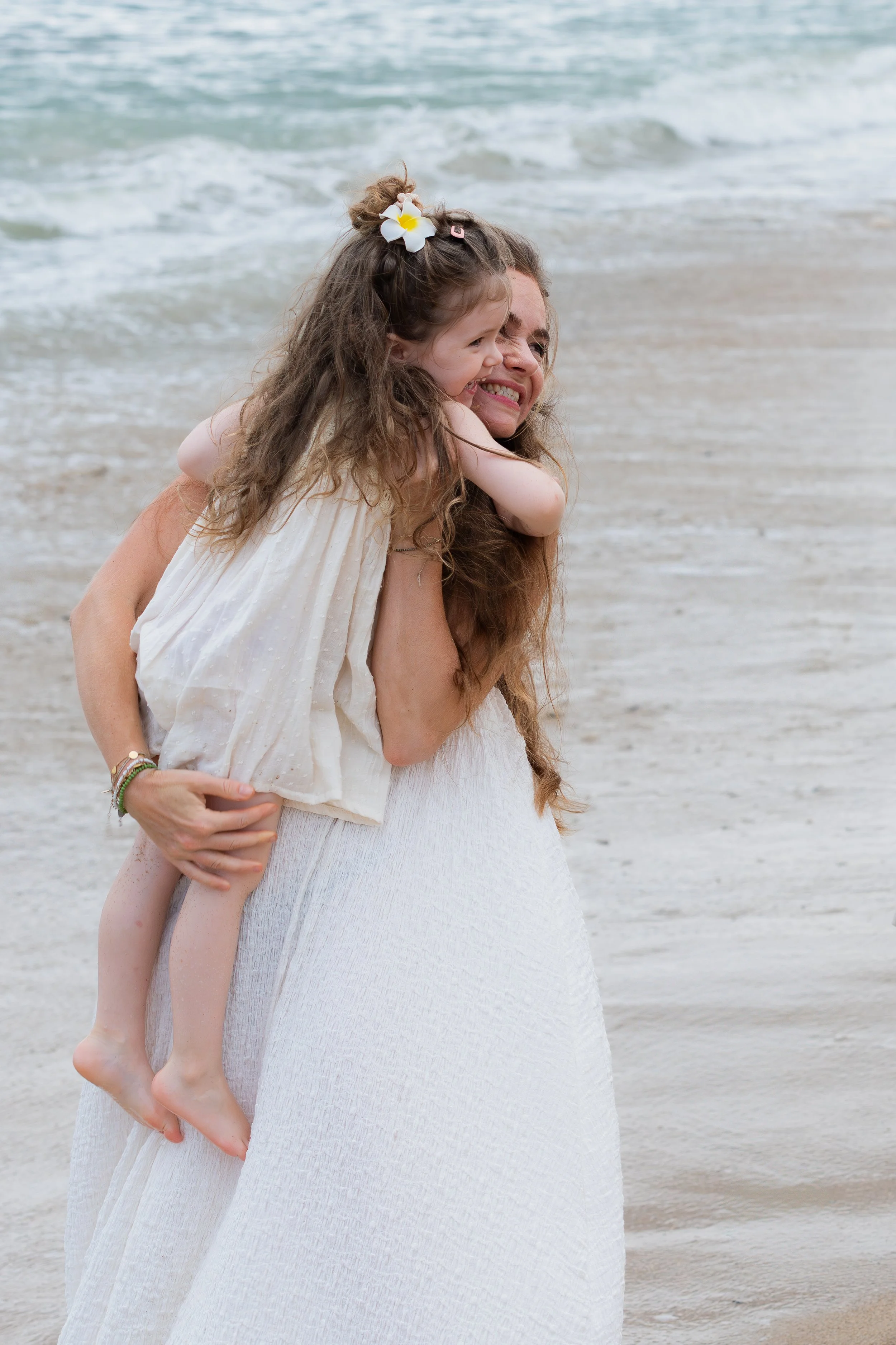 Een vrouw draagt een jonge meisje op haar schouders op het strand, beiden lachen en genieten van de dag.