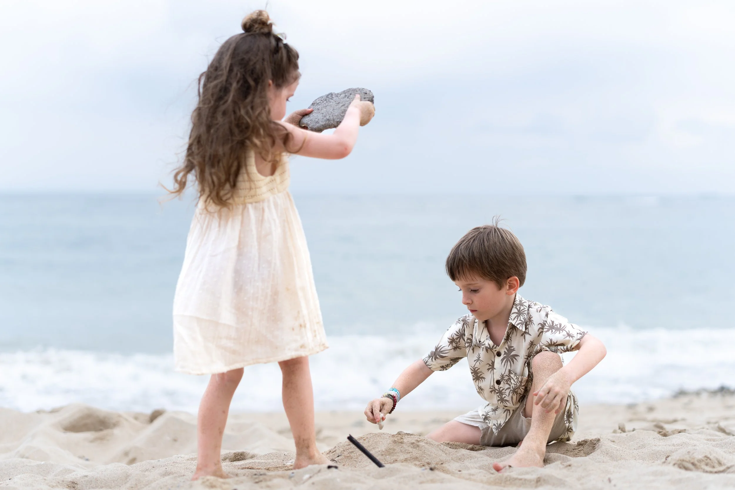 Twee kinderen spelen op het strand, de meisjes met een steen en de jongen met zand en een schepje.