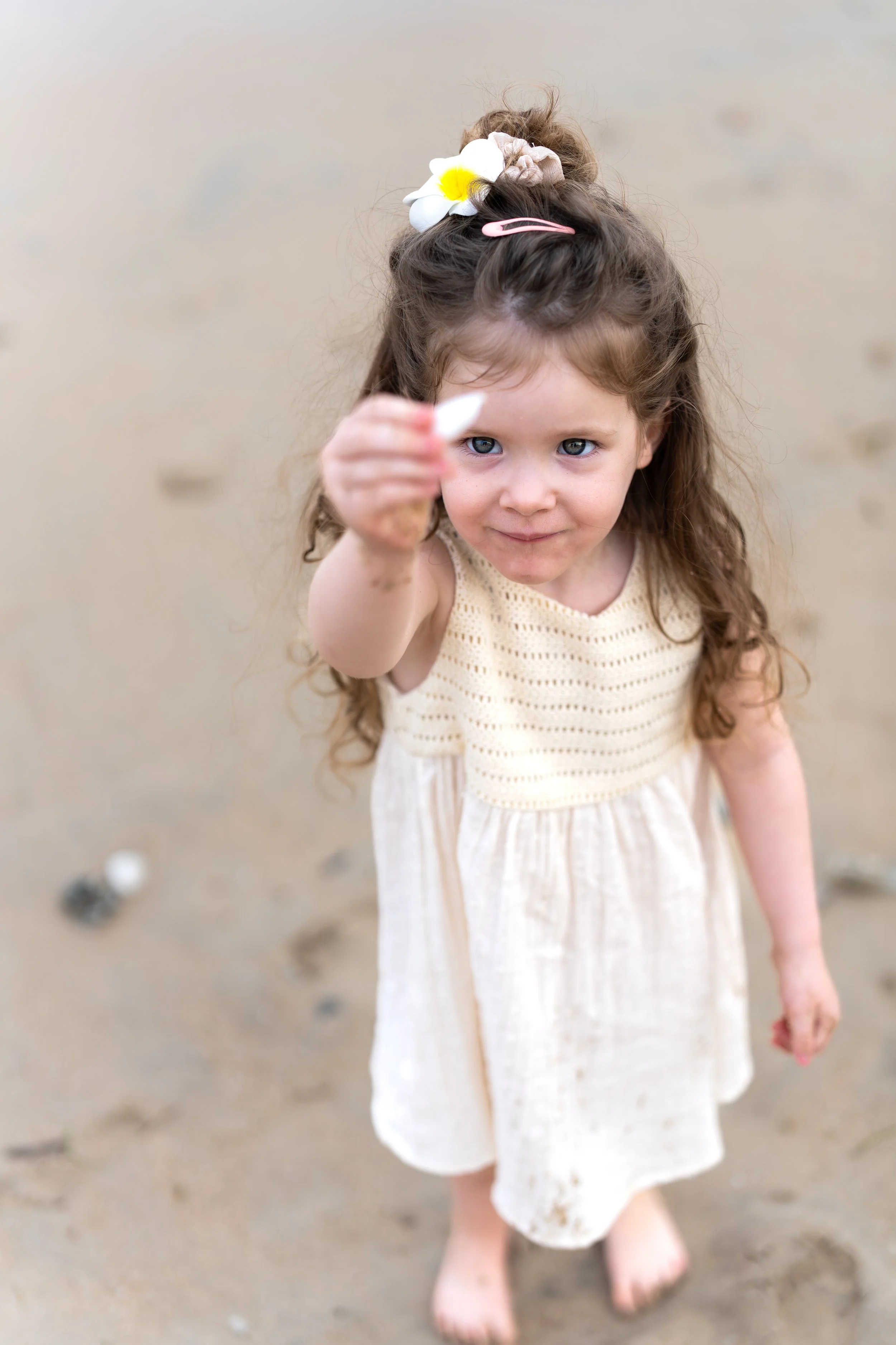 Een jong meisje met lang, krullend bruin haar, gekleed in een lichte jurk, staat op het zand van het strand en houdt een wit schelpje naar de camera, met een speelse blik en een bloem in haar haar.