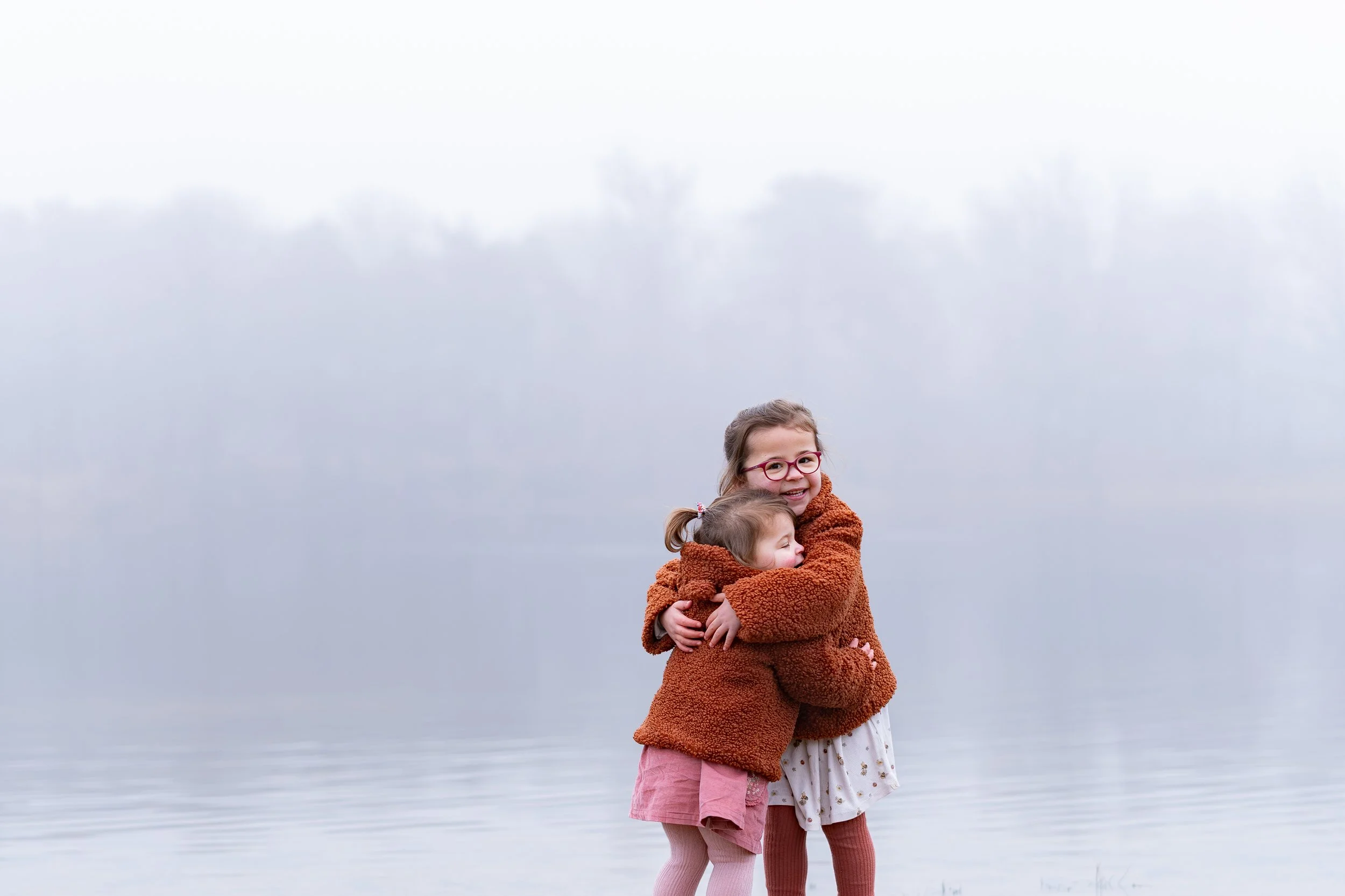 Twee jonge meisjes in herfstkleding die elkaar omhelzen aan de rand van een water, met mistige achtergrond en wind}