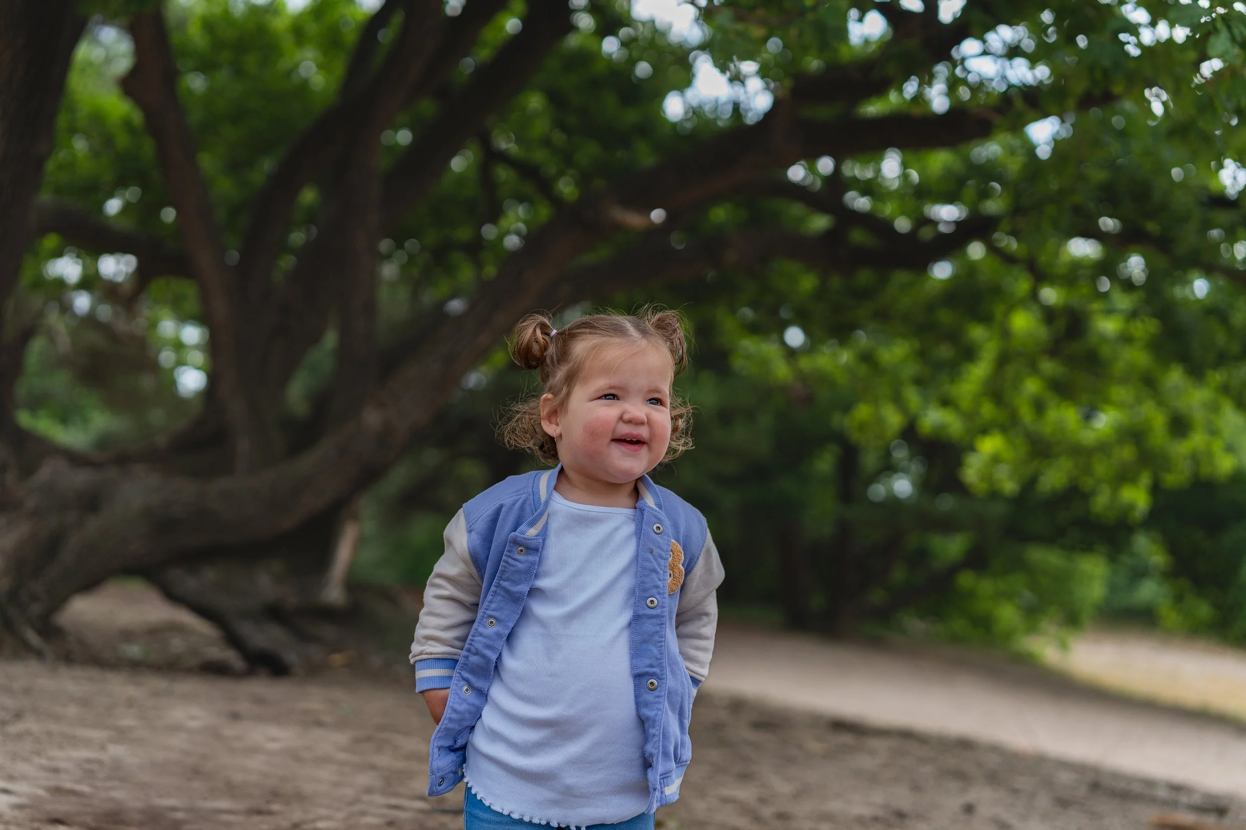 Kleine meisjesbaby met krullend haar in een blauwe jas en witte T-shirt, lachend in een bos met grote bomen en groene bladeren.