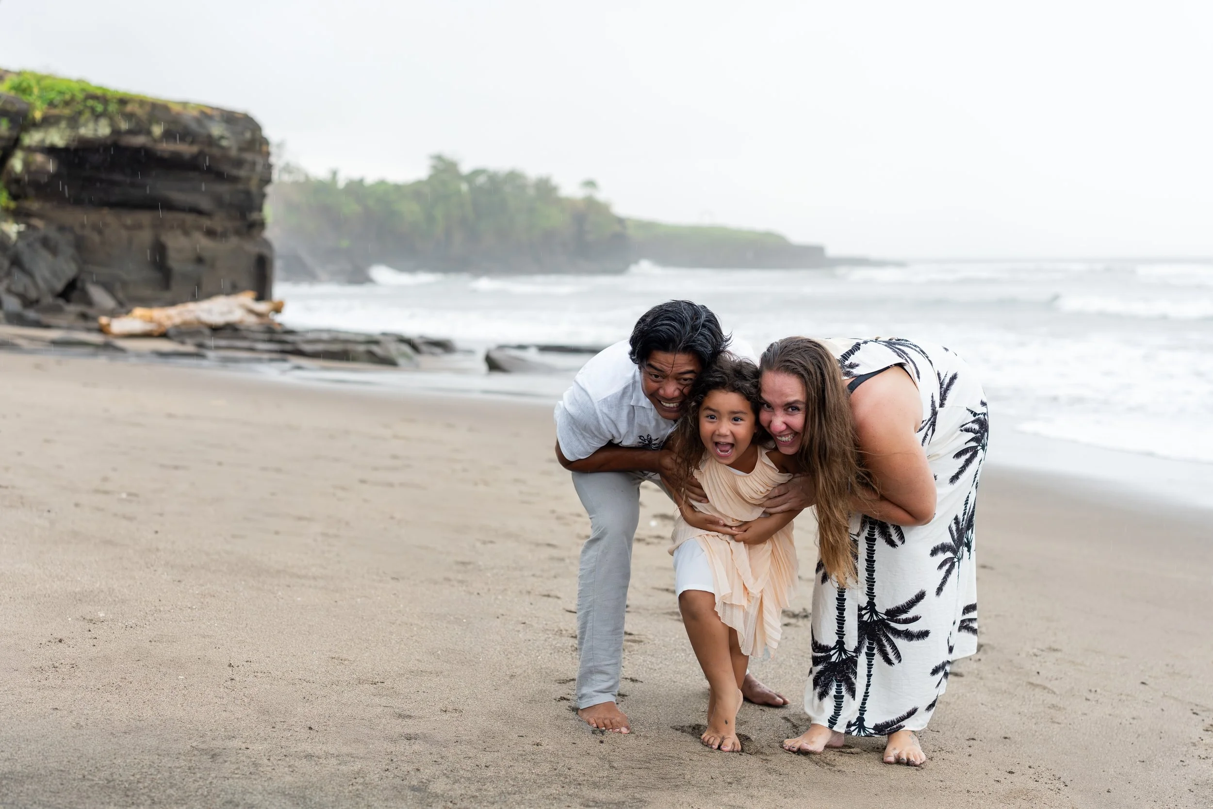 Een gezin van drie mensen, een man, een vrouw en een jong meisje, lachen en knuffelen op het strand tijdens een regenachtige dag.