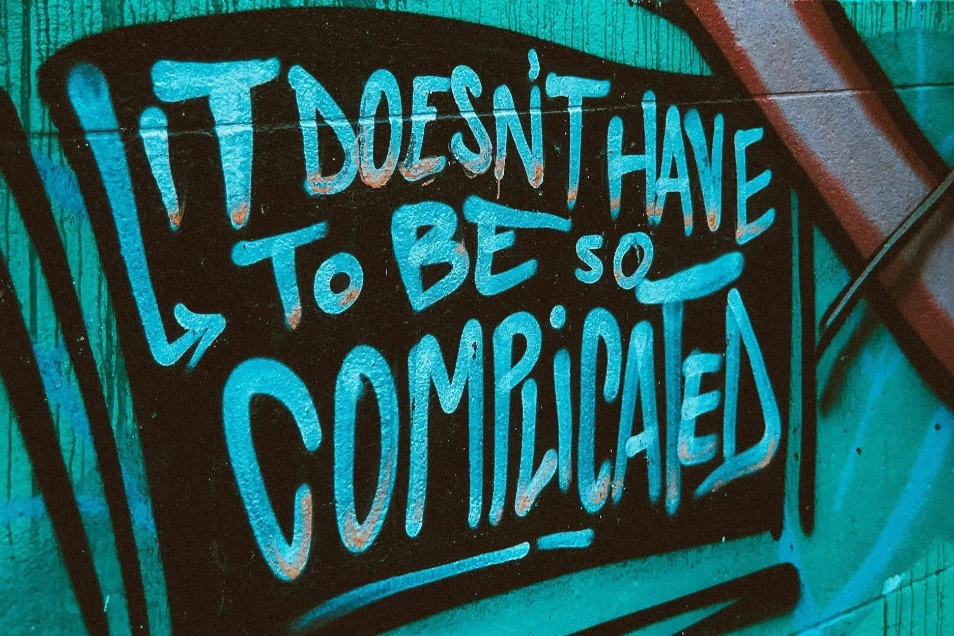 A graffiti-style mural on a wall with the quote, 'It doesn't have to be so complicated,' written in blue and black spray paint with a teal background.