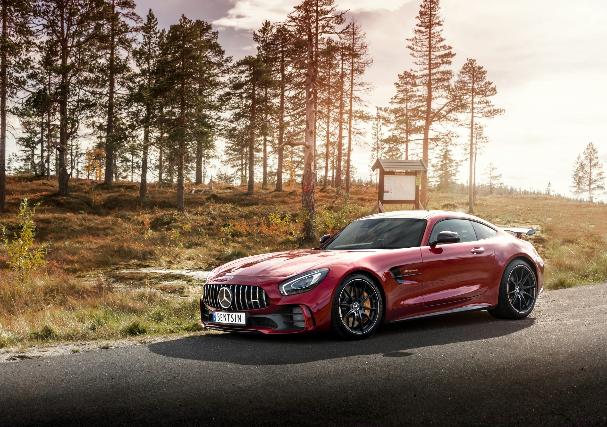 A red Mercedes-Benz sports car with a license plate reading BENTSIN is parked on the side of a rural road, with a forest and an informational sign in the background during sunset.