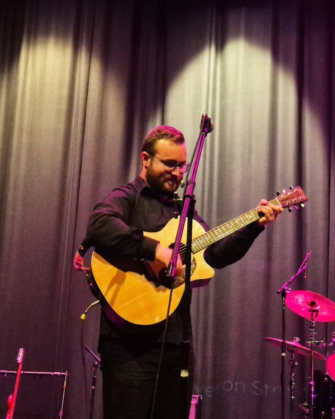 A man playing an acoustic guitar on stage, smiling with gray curtains in the background, microphone stand in front.