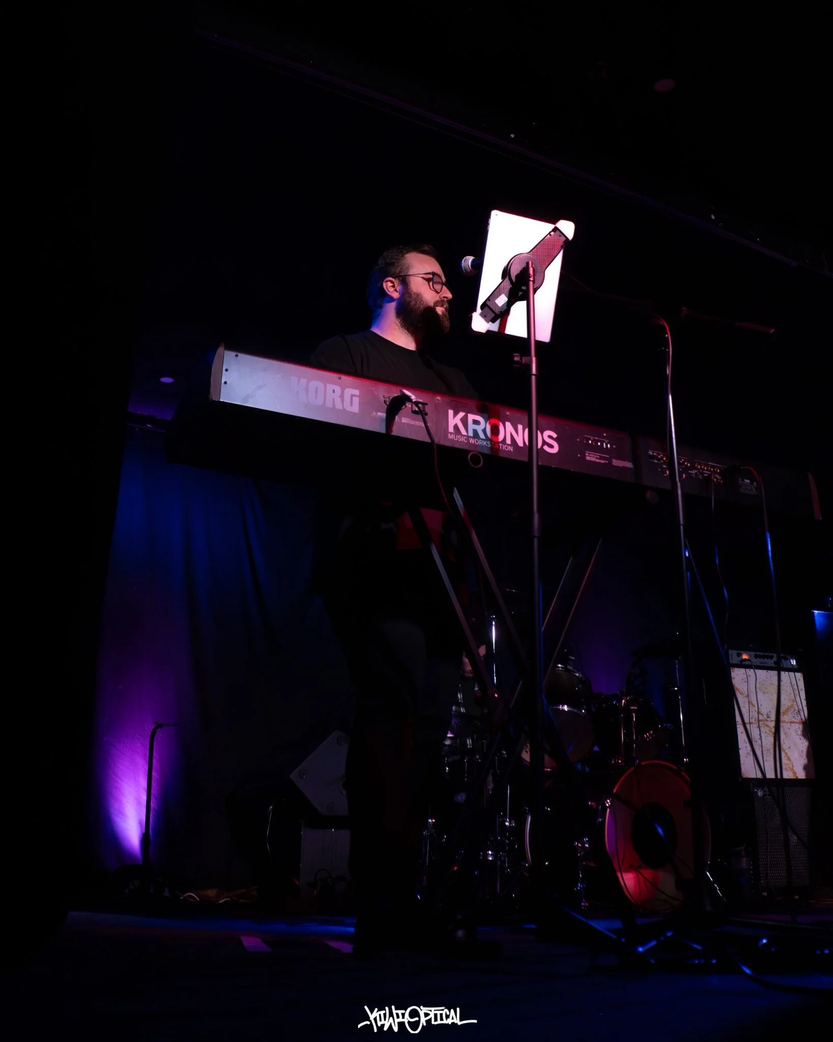 A musician with a beard, glasses, and headphones, playing a Korg Kronos keyboard on a dark stage, illuminated by purple and blue lights, with a sheet music stand in front of him.