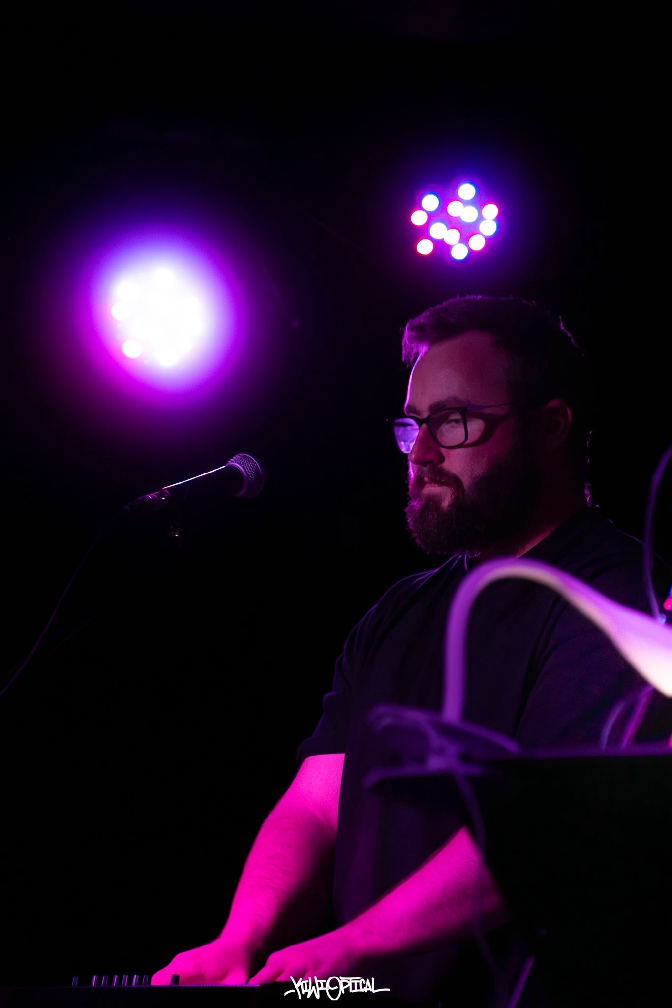 A man with glasses and a beard playing keyboard on stage with purple and blue stage lights behind him.