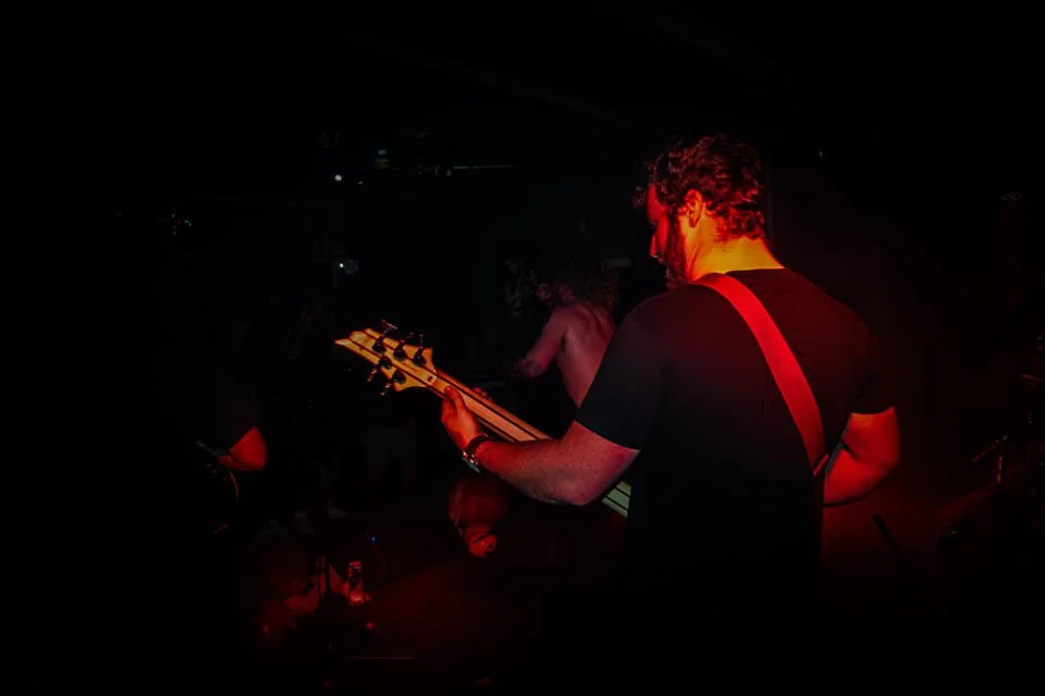 Man playing electric guitar on stage in a dimly lit concert or club setting.