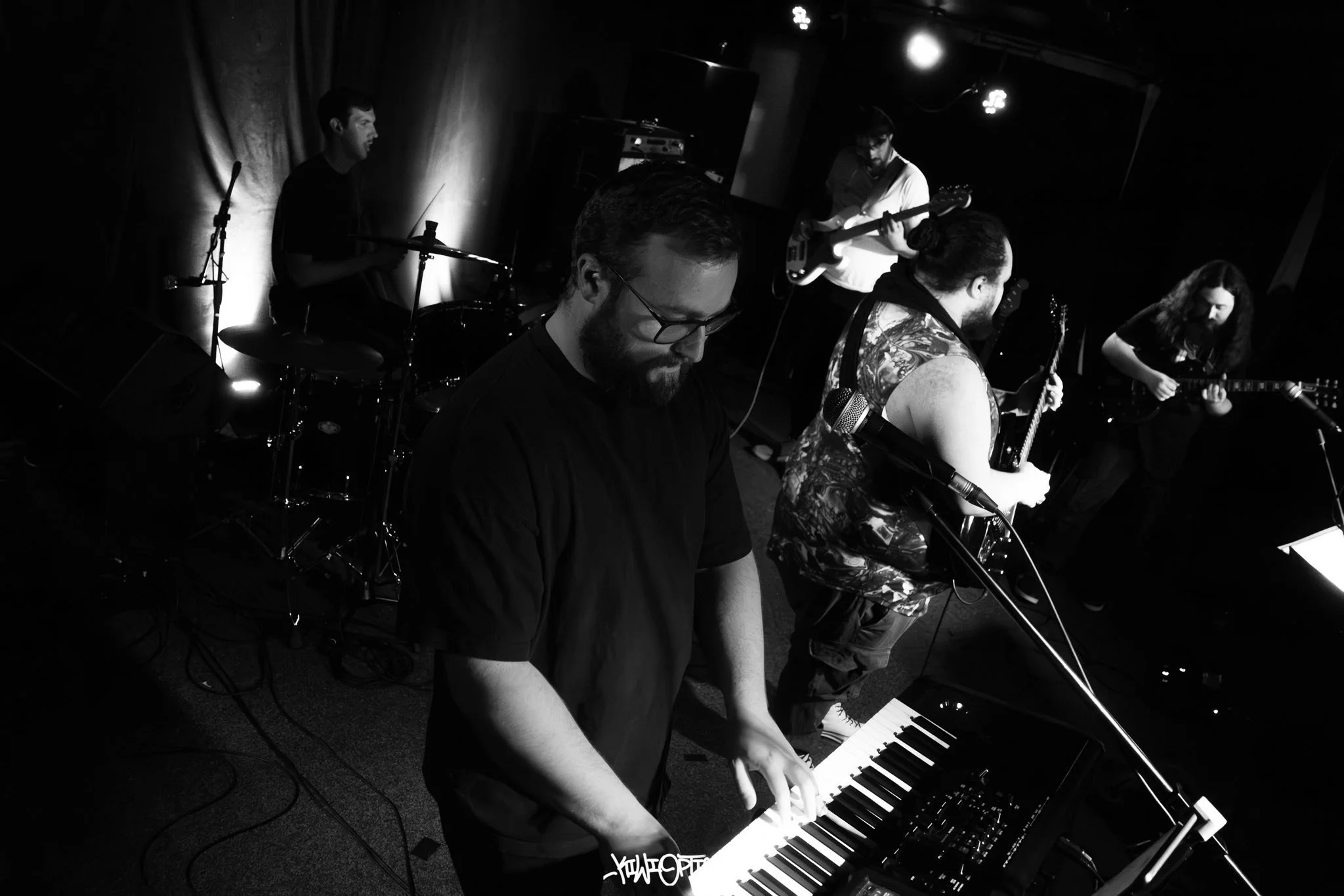 A black and white photo of a band performing live on stage, with a pianist in the foreground, a drummer in the back, and two guitarists to the right.
