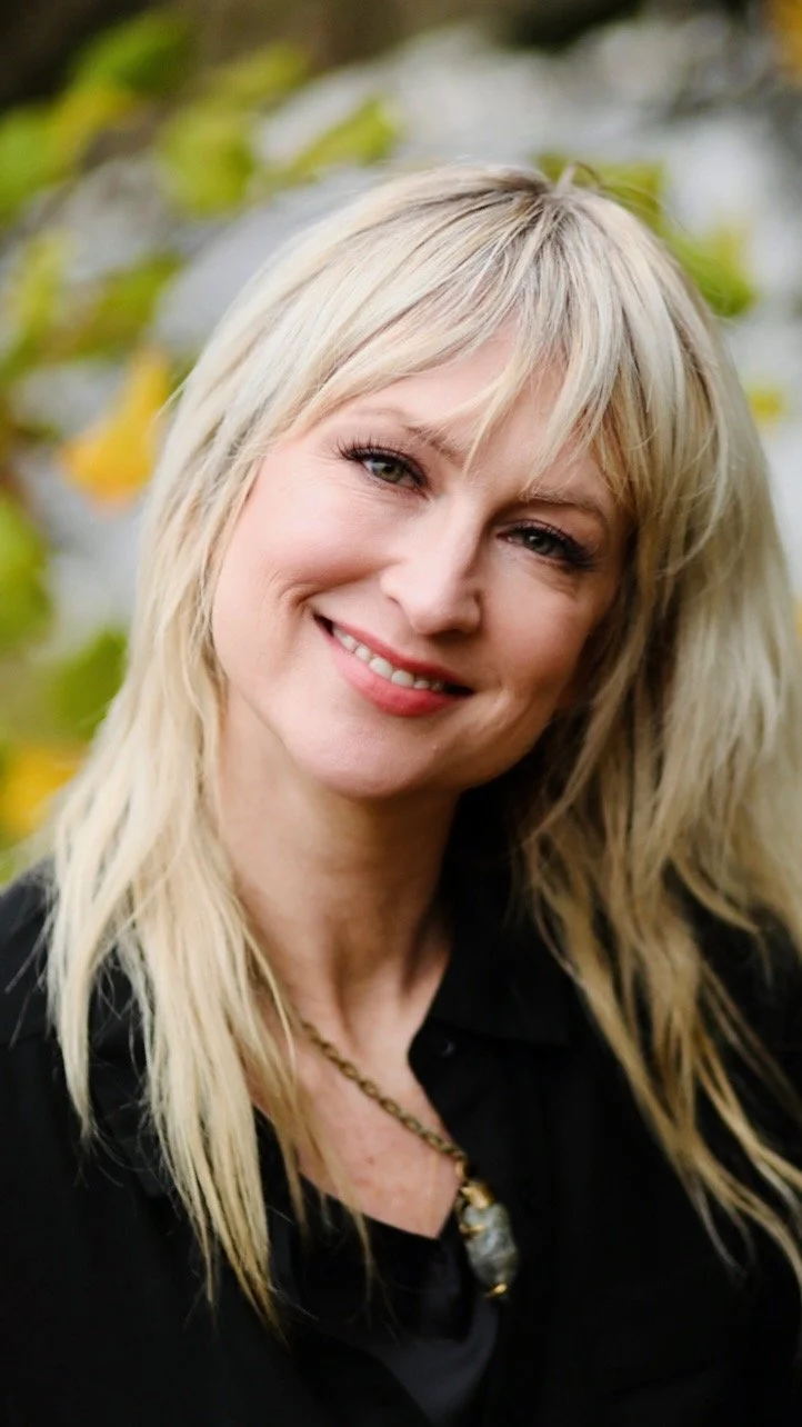 Rebecca, smiling outdoors with green foliage in the background.