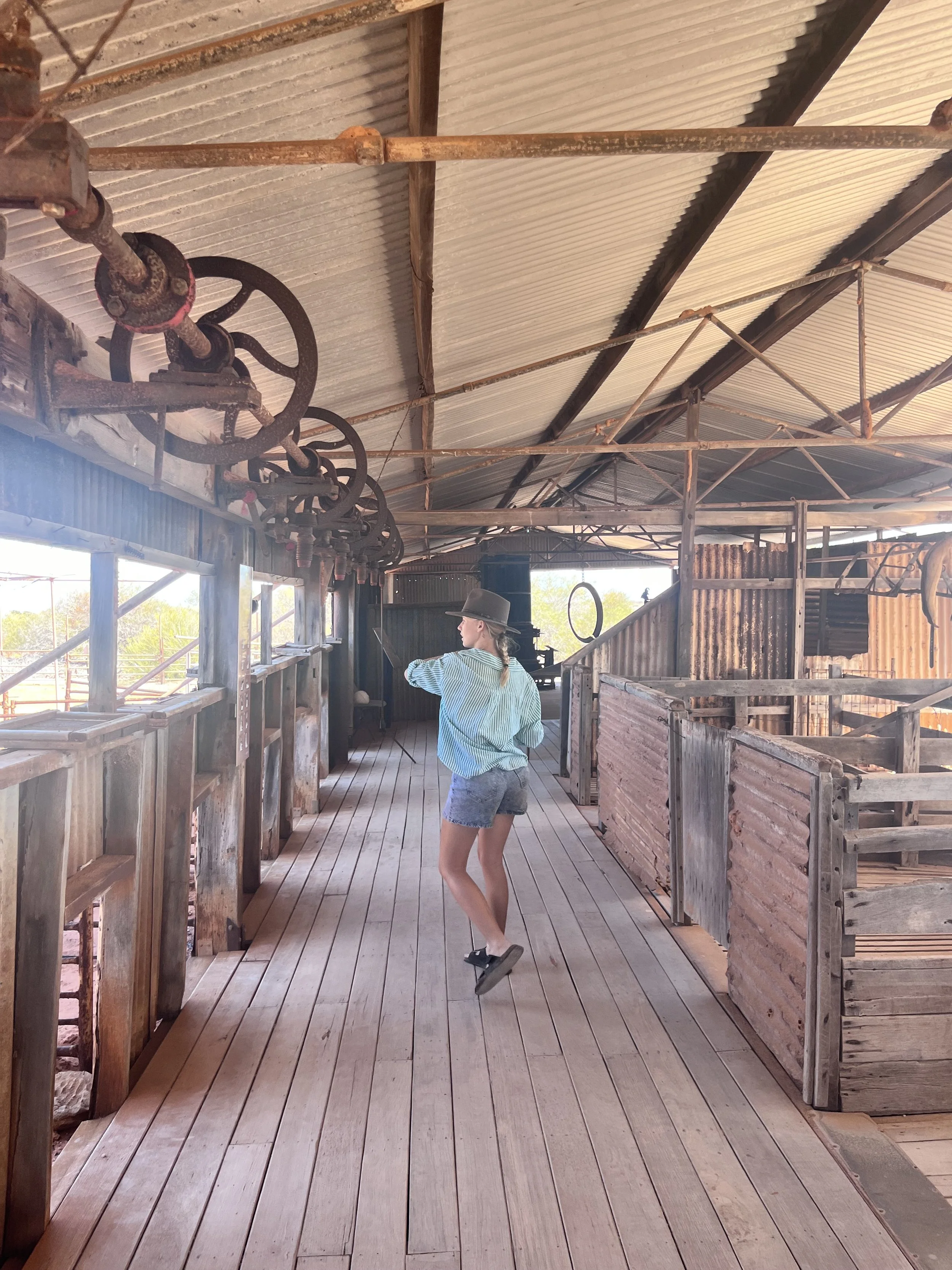 A woman wearing a hat, striped shirt, and denim shorts standing on a wooden platform inside a rustic barn, pointing toward something, with farming equipment hanging from the ceiling.