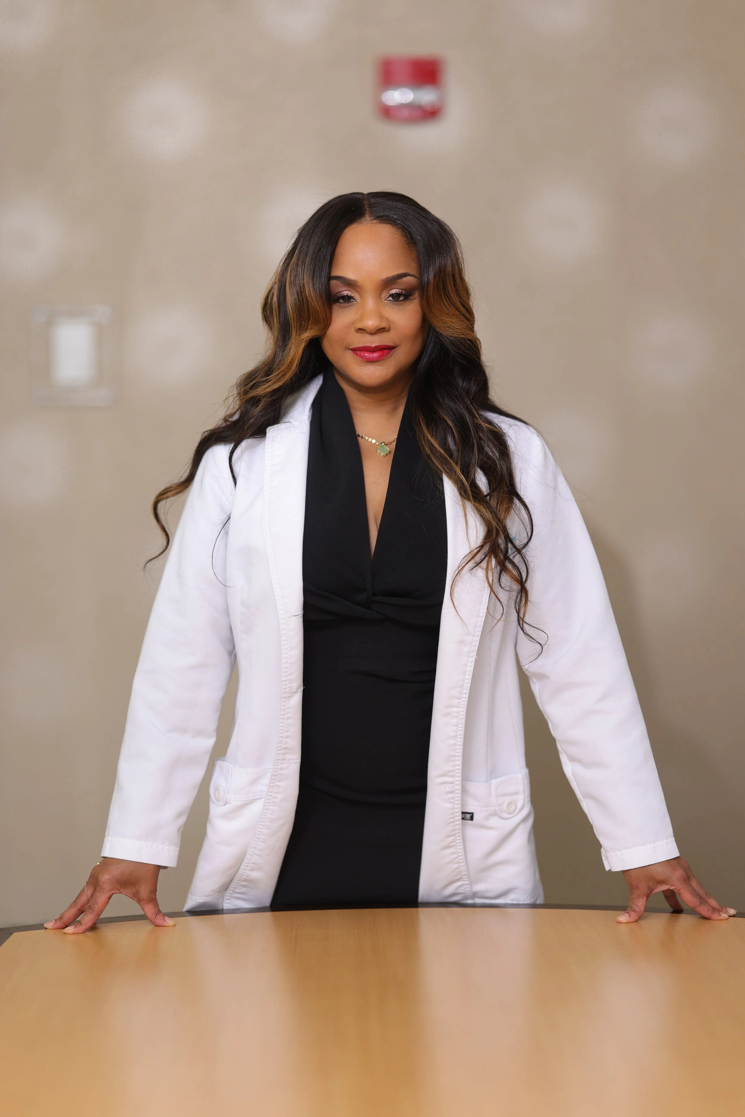 A woman wearing a white lab coat standing behind a wooden table with her hands resting on it, in a room with beige walls and a red fire alarm on the wall behind her.