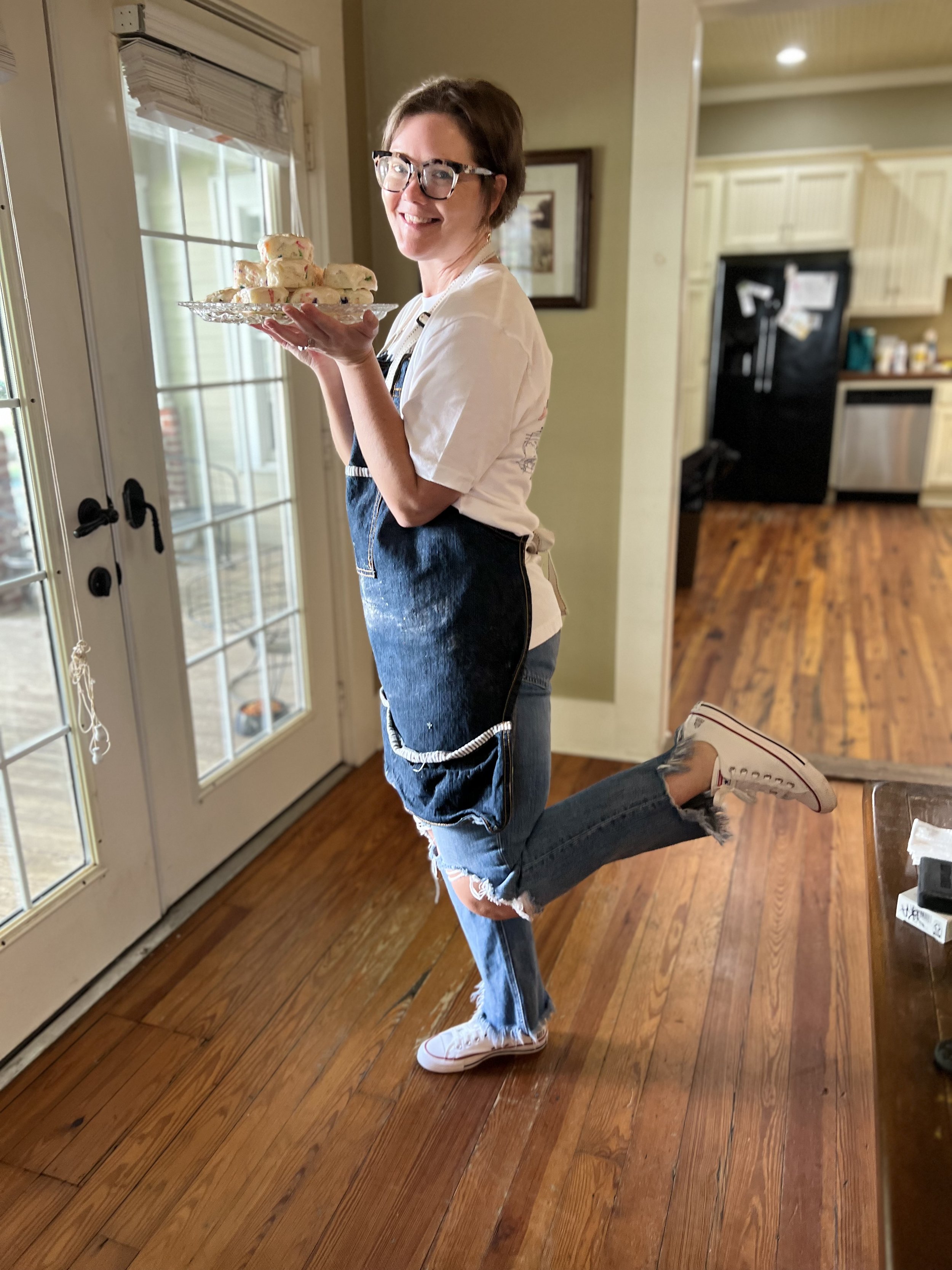 A woman with short brown hair, glasses, a white T-shirt, ripped jeans, and white sneakers is holding a plate with cookies, standing inside a kitchen near a glass door.