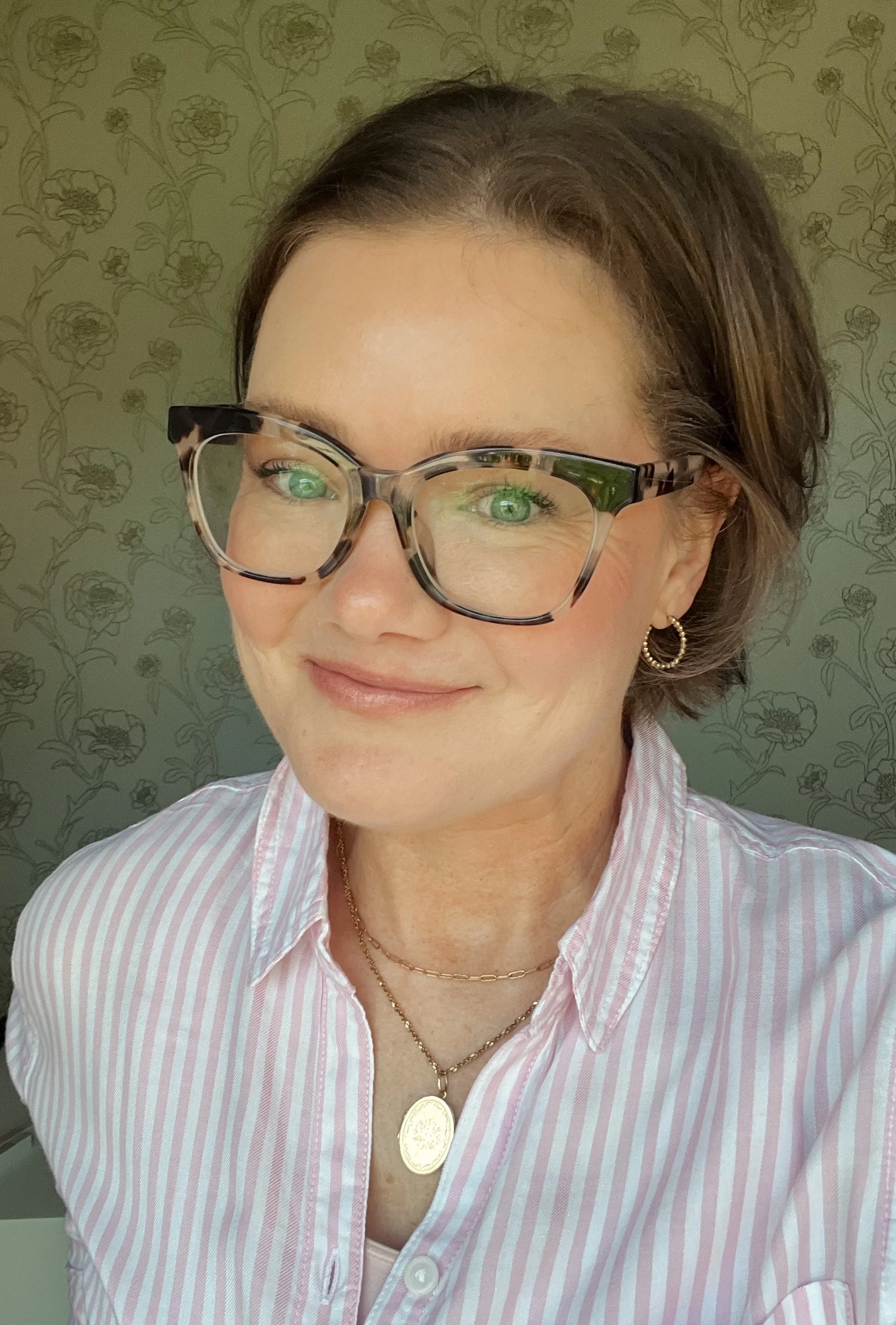 A woman with short brown hair wearing large tortoiseshell glasses, gold hoop earrings, and layered gold necklaces, smiling while looking slightly to her right. She is dressed in a pink and white striped button-up shirt, standing in front of a floral wallpaper background.