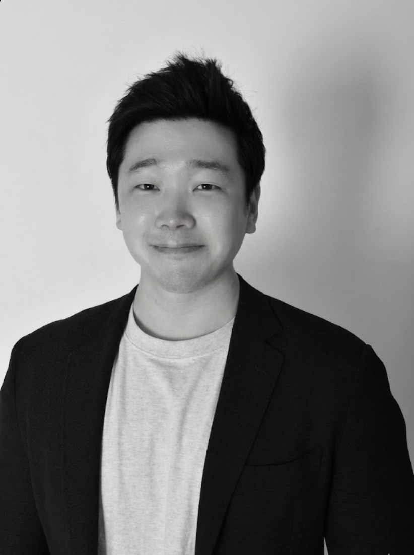 Black and white portrait of a smiling young Asian man with short, styled hair wearing a light-colored T-shirt and a dark blazer against a plain background.