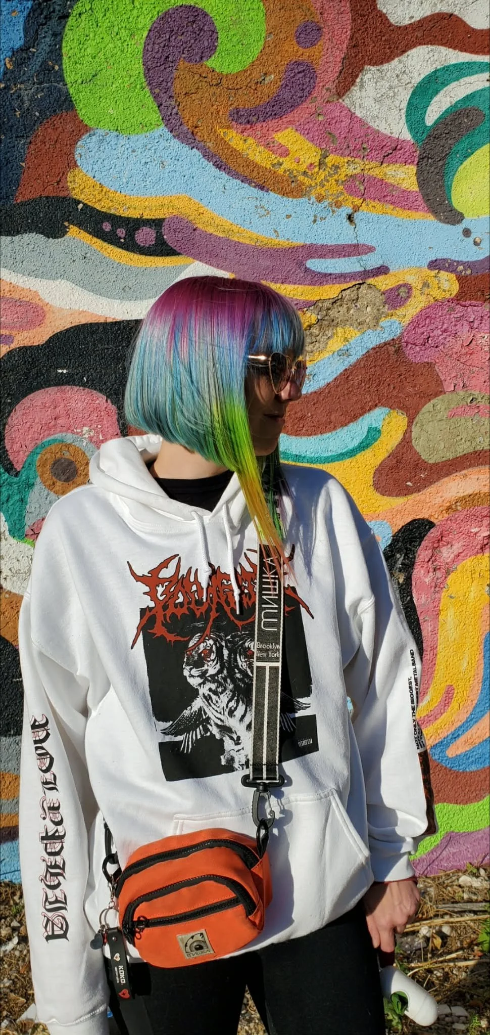 A woman with shoulder-length, rainbow-colored hair, wearing sunglasses and a white hoodie with black and red graphics, standing against a vibrant graffiti mural with swirling colorful patterns.