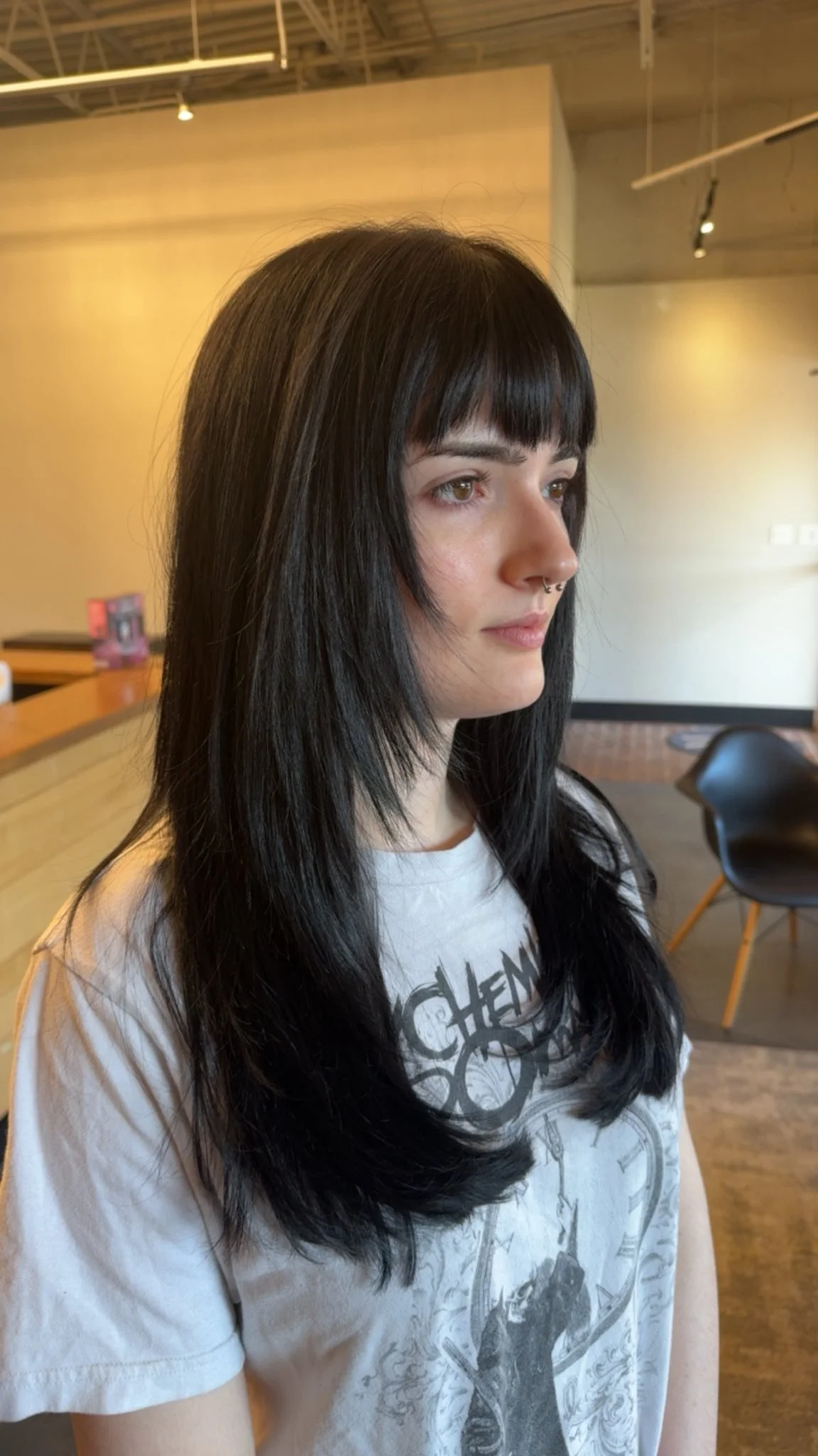 A young woman with long black hair and a septum piercing looking to the right, in a modern indoor space with wooden and concrete decor.