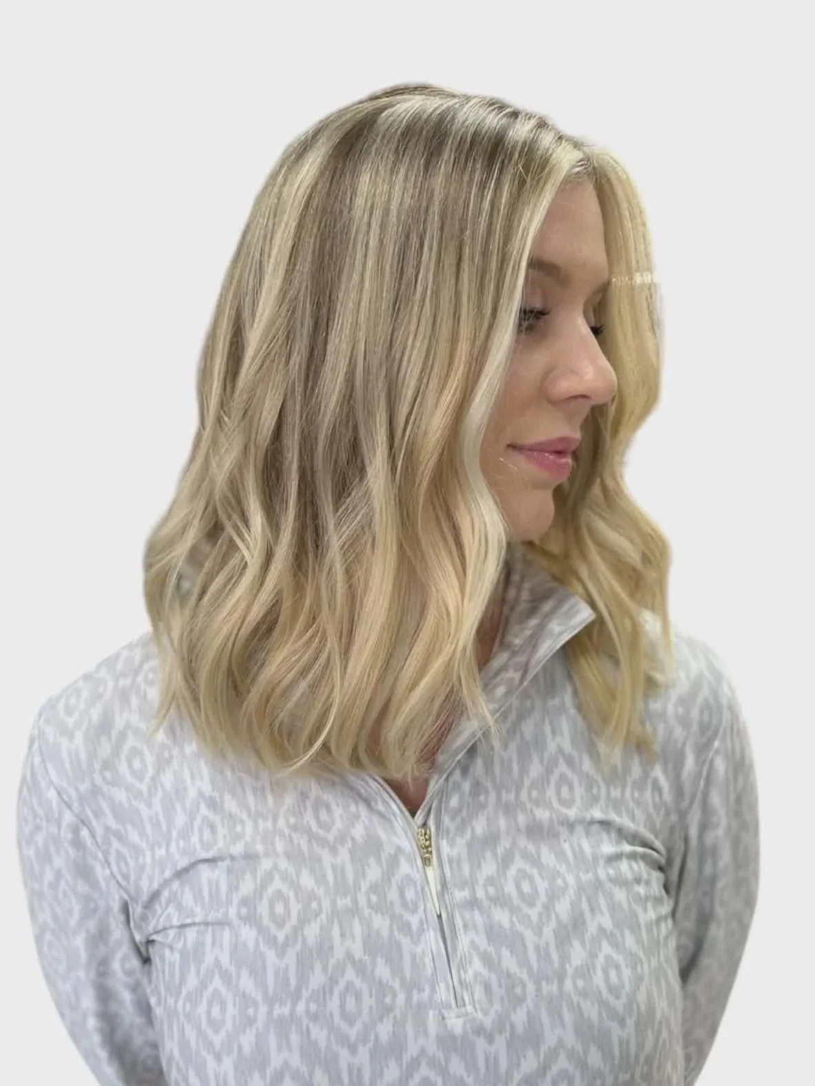 A woman with blonde, wavy hair and light skin, wearing a light gray zip-up jacket with a subtle pattern, looking down and to the side against a plain background.