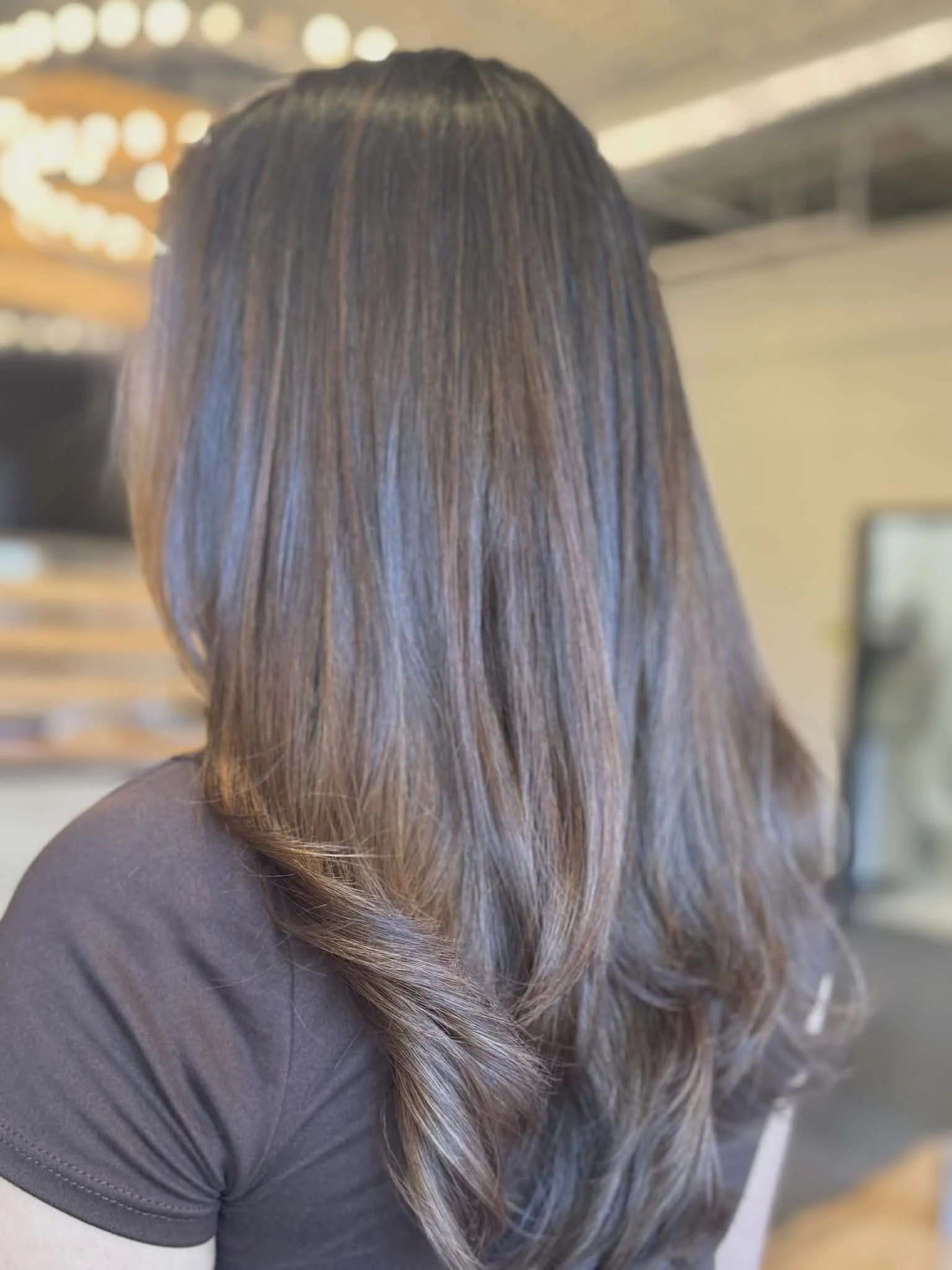 Back view of a woman with medium length, brown, wavy hair, wearing a dark gray shirt, in an indoor setting.