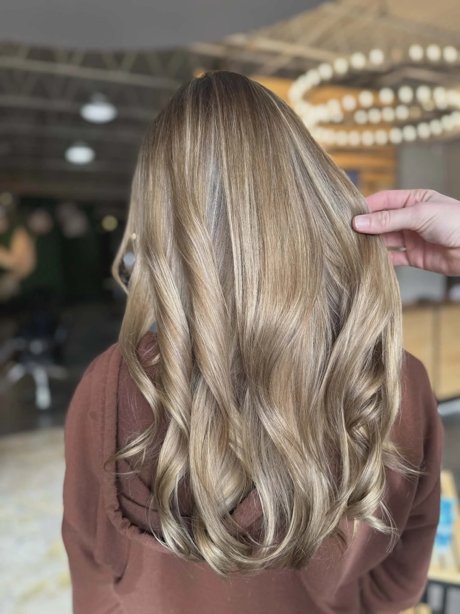 Long blonde wavy hair on a person wearing a brown top, held by a hand on the side, in a salon or studio setting.