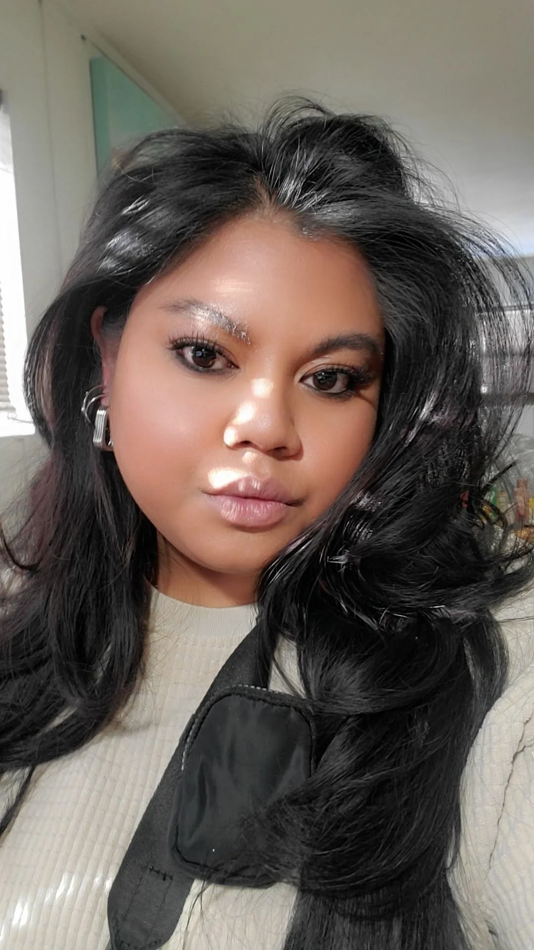 A woman with long black hair and earrings, looking at the camera indoors with natural light.